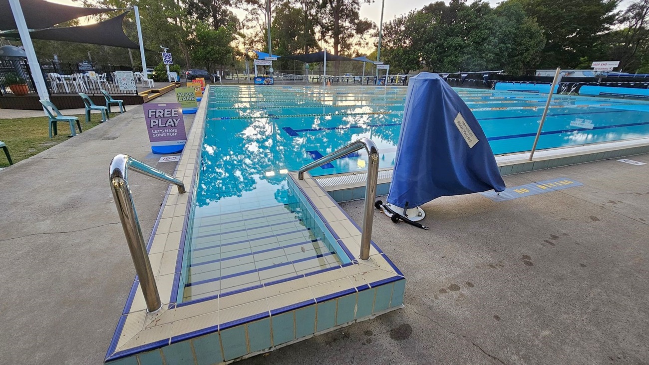 A swimming pool with clear blue water and lane dividers. In the foreground, metal handrails lead into the pool. On the right, a covered piece of equipment is on the pool deck. Signs along the poolside read "FREE PLAY" and "SLOW LANE." To the left, there are green plastic chairs and a shaded area with trees in the background. The pool deck is concrete with some wet patches.