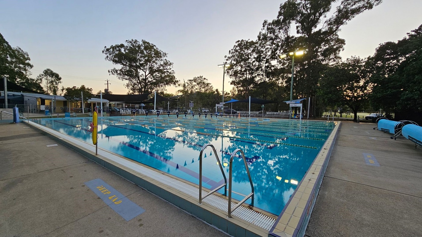 An outdoor swimming pool with clear water reflecting nearby trees. On the left, there are buildings and a few chairs. The pool has lane dividers and a metal ladder in the foreground. On the right, there are blue pool covers on rollers and more trees. The surrounding area is paved with concrete, and a sign on the ground reads "DEEP END." Overhead lights are visible, and the sky is transitioning to dusk.