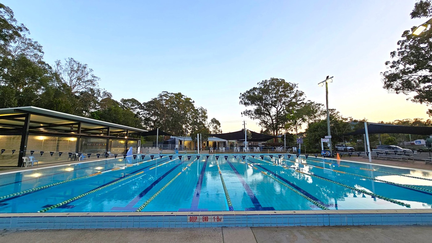 An outdoor swimming pool with multiple lanes, marked by blue and yellow lane dividers. On the left, there's a covered seating area with chairs. In the background, trees and a few buildings are visible. On the right, there are more chairs and a couple of parked cars. The pool's edge is labelled "DEEP END 1.8m". The area is lit by tall lights, and the sky is clear.