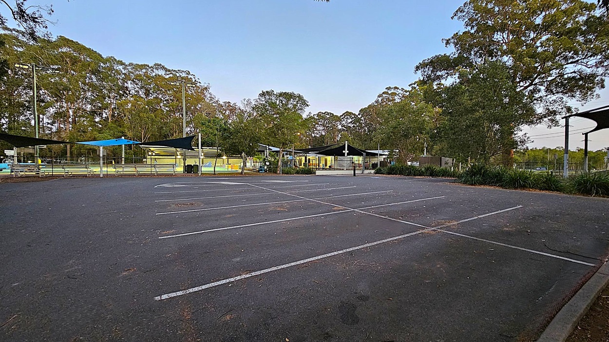 A parking area with a marked disabled parking space on the right. In the centre, a black bollard with red reflectors stands on a striped section of the pavement. Behind, a fenced area with a playground is visible, featuring a shade structure and play equipment. Trees and greenery surround the area, with a palm tree on the left and dense foliage on the right. The ground is covered with scattered leaves.