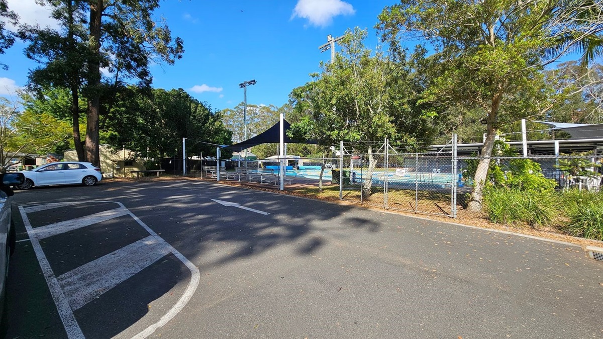 A car park with a white car on the left. The road has white painted lines and an arrow. On the right, there's a fenced area with a swimming pool covered by a black shade sail. Trees and greenery surround the area, with tall trees in the background. The ground is paved, and there are shadows from the trees.