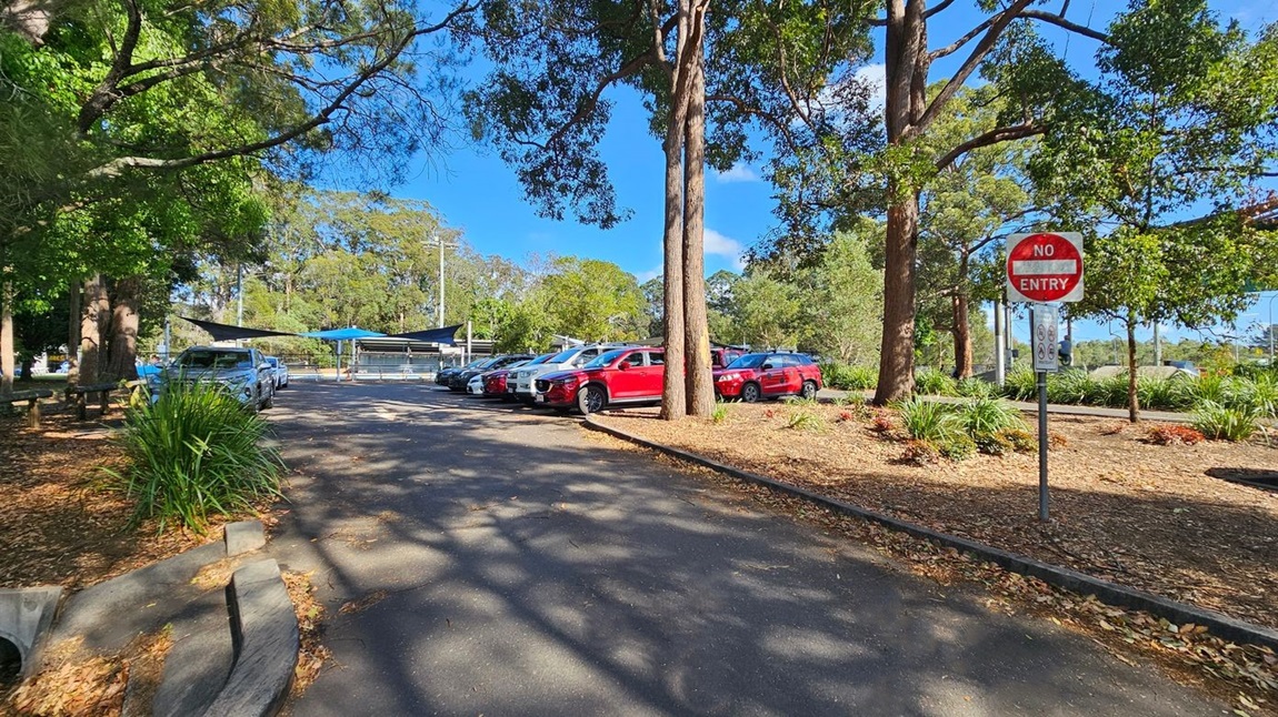 A car park with several vehicles, including red and white cars, is visible on the right. A "No Entry" sign is prominently displayed near the parked cars. Trees with green foliage surround the area, casting shadows on the ground. On the left, there are benches and a patch of grass with fallen leaves. In the background, a structure with blue shade sails is partially visible, surrounded by more trees.