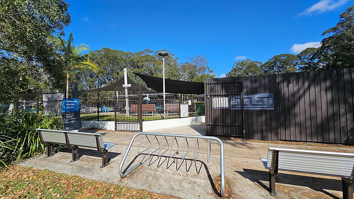 A wooden fence with the sign "Sunshine Coast Council Beerwah Aquatic Centre" is on the left, surrounded by lush green bushes. A concrete path leads to the right, where people are walking towards a blue sign. Tall trees and power lines are visible in the background. A bench is placed near the fence.