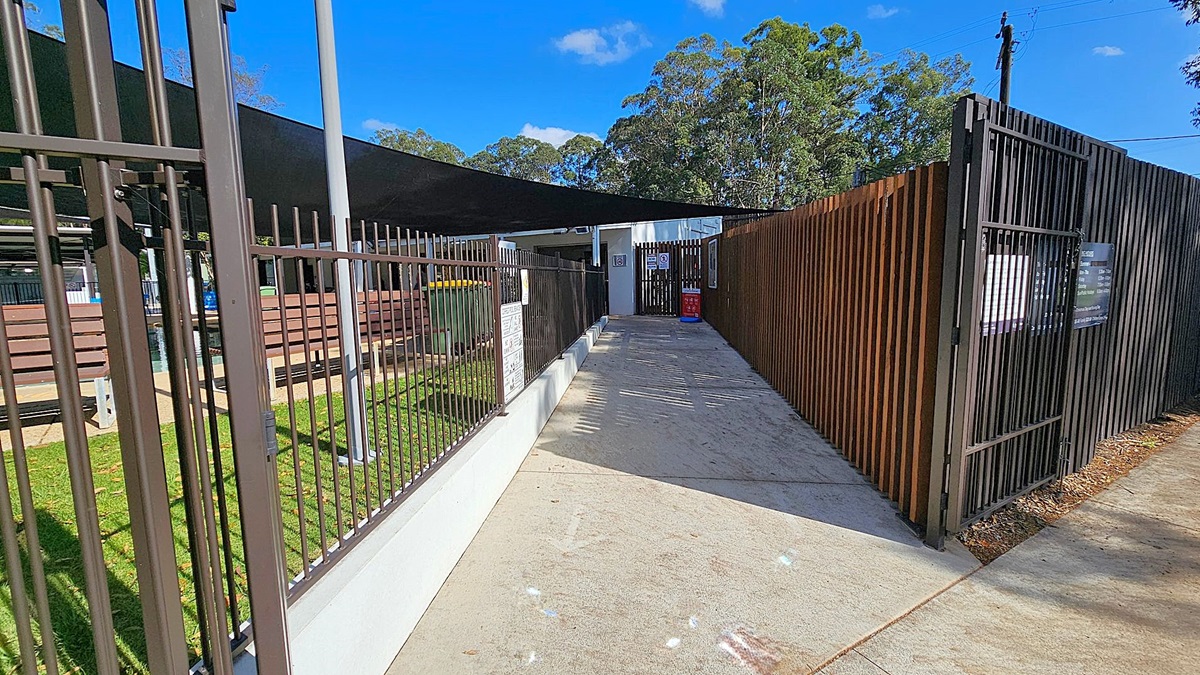 A pathway with a concrete surface leads to a gated entrance. On the left, there's a metal fence with a grassy area and benches visible behind it. On the right, a wooden slat fence runs alongside the path. Overhead, a black shade cloth is stretched across, providing partial cover. In the background, there are trees and a building with a sign near the entrance. The sky is clear and blue.