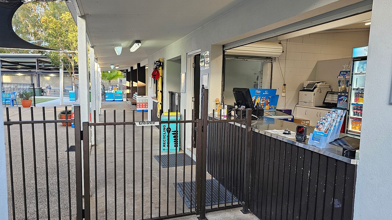 A gated entrance with a black metal fence leads to a covered area. On the right, there's a counter with a computer, brochures, and a fridge stocked with drinks. The ceiling has fluorescent lights. In the background, outdoor seating with blue chairs and potted plants is visible, along with a recycling bin. Trees are seen beyond the seating area.