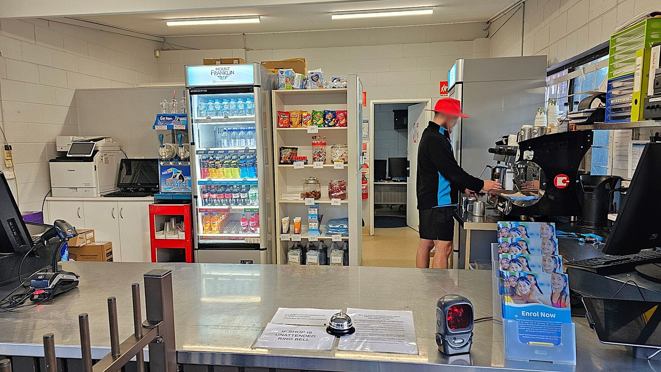 A small shop interior with a counter in the foreground, featuring a bell and a card reader. Behind the counter, a person in a red hat operates a coffee machine. To the left, a fridge stocked with drinks and a shelf displaying snacks and other items. A printer and computer are on a side counter. The walls are white, and the ceiling has fluorescent lights. A doorway in the background leads to another room.