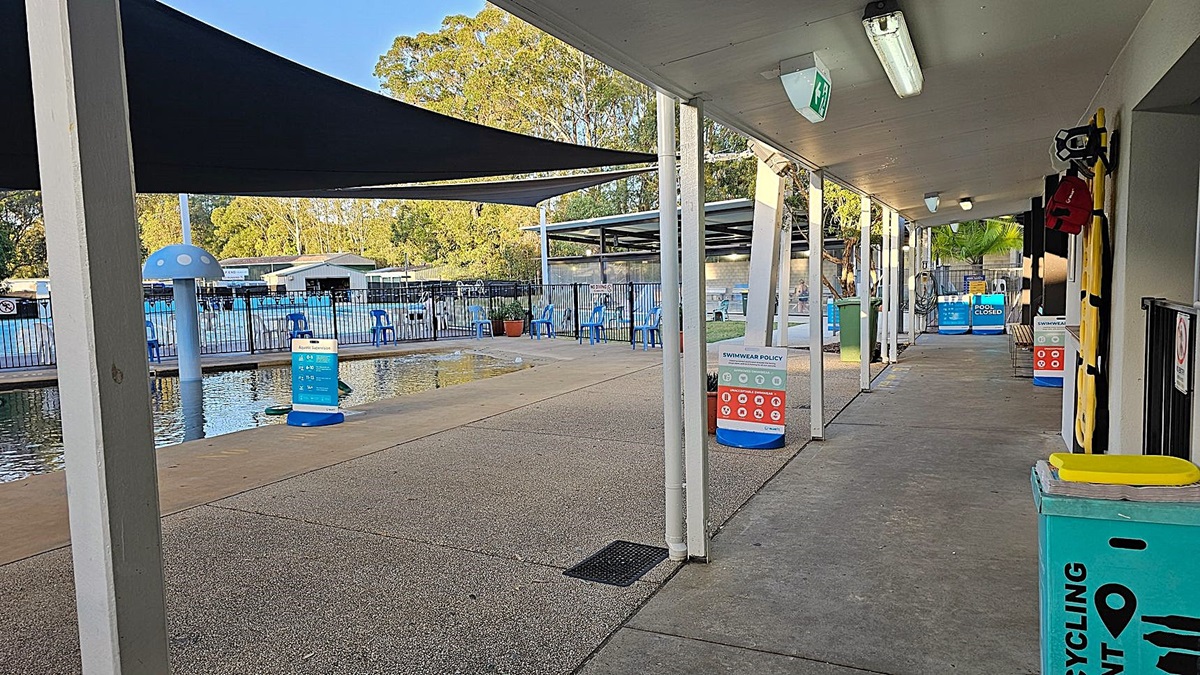 A pool area with a shaded walkway on the right, featuring a recycling bin and swimwear policy signs. The walkway has a concrete floor and a ceiling with fluorescent lights. On the left, there's a fenced pool with blue chairs and a mushroom-shaped water feature. In the background, trees and a building are visible.