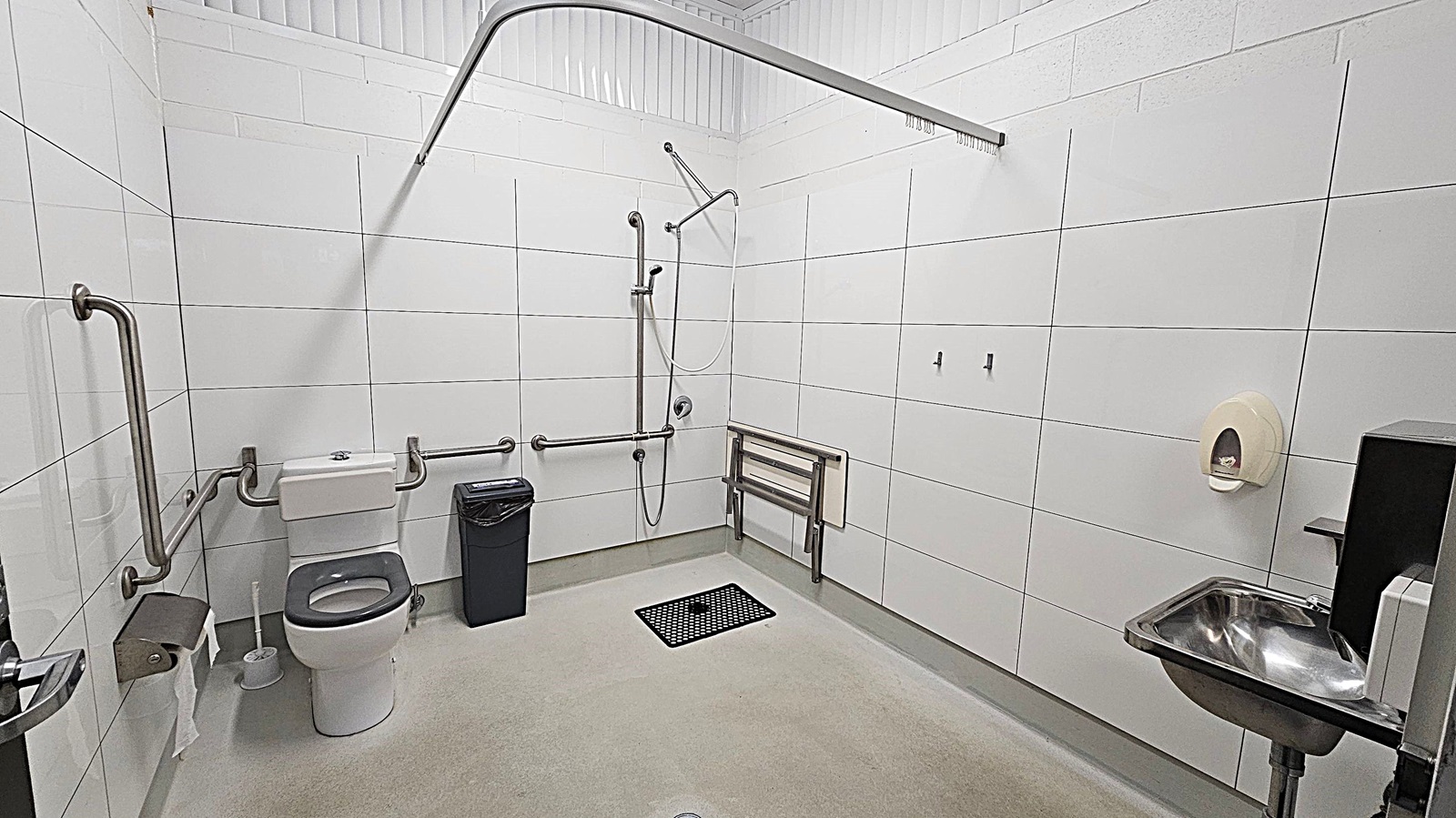 A spacious, accessible bathroom with white tiled walls and a light green floor. On the left, there is a toilet with grab bars and a toilet paper holder. A black bin is next to the toilet. In the centre, a shower area with a handheld showerhead and additional grab bars. On the right, a foldable shower seat is mounted on the wall. A stainless steel sink with a soap dispenser is also on the right. The ceiling is white with a curtain rail above the shower area.