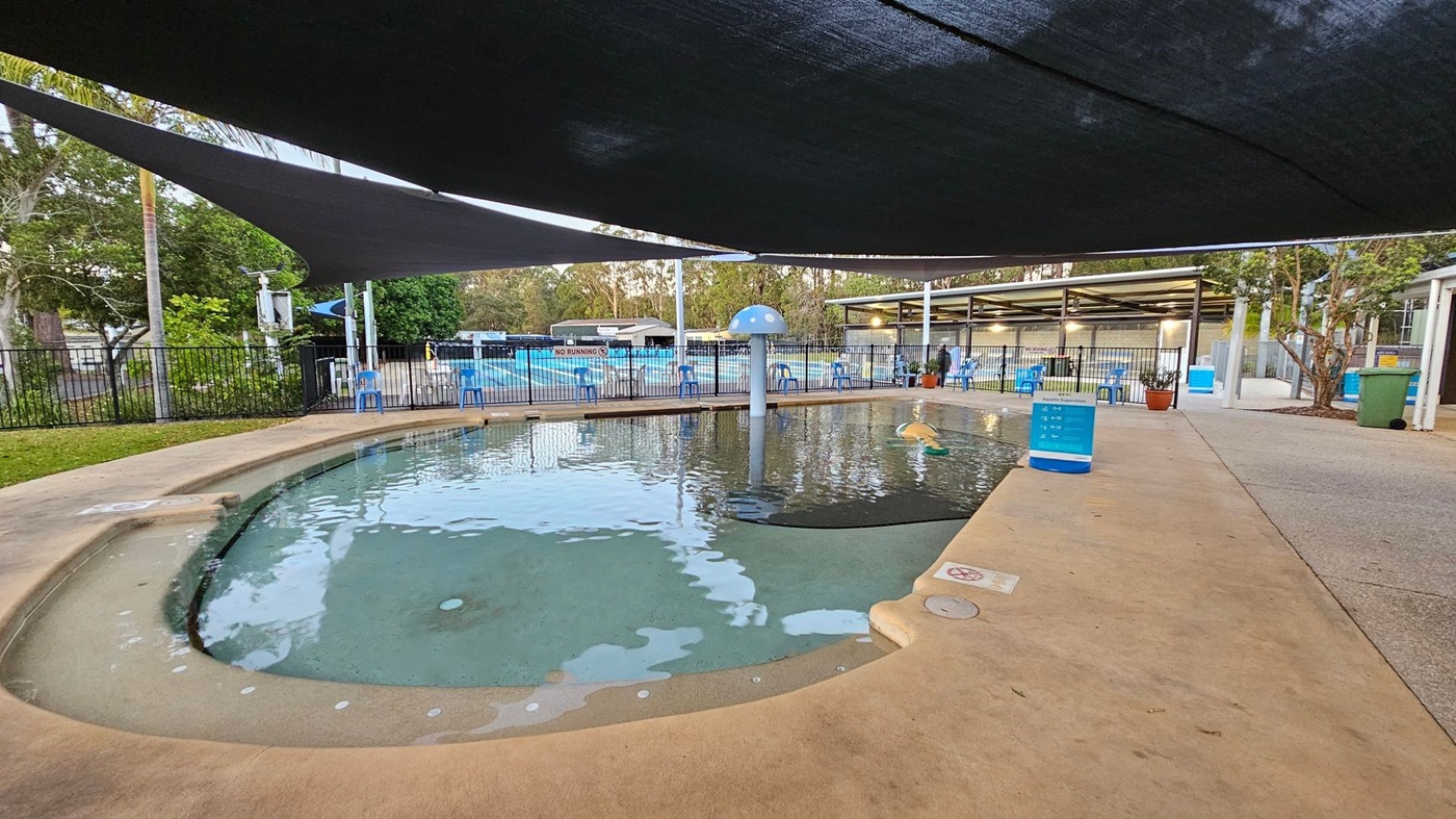A shallow children's pool with a mushroom-shaped water feature and a floating toy. The pool is surrounded by a concrete walkway and enclosed by a black metal fence. Blue plastic chairs are placed along the fence. Overhead, there are large shade sails. In the background, there are trees, a few parked cars, and a wooden fence. Signs indicate pool rules, and there are bins and a bench near the entrance.