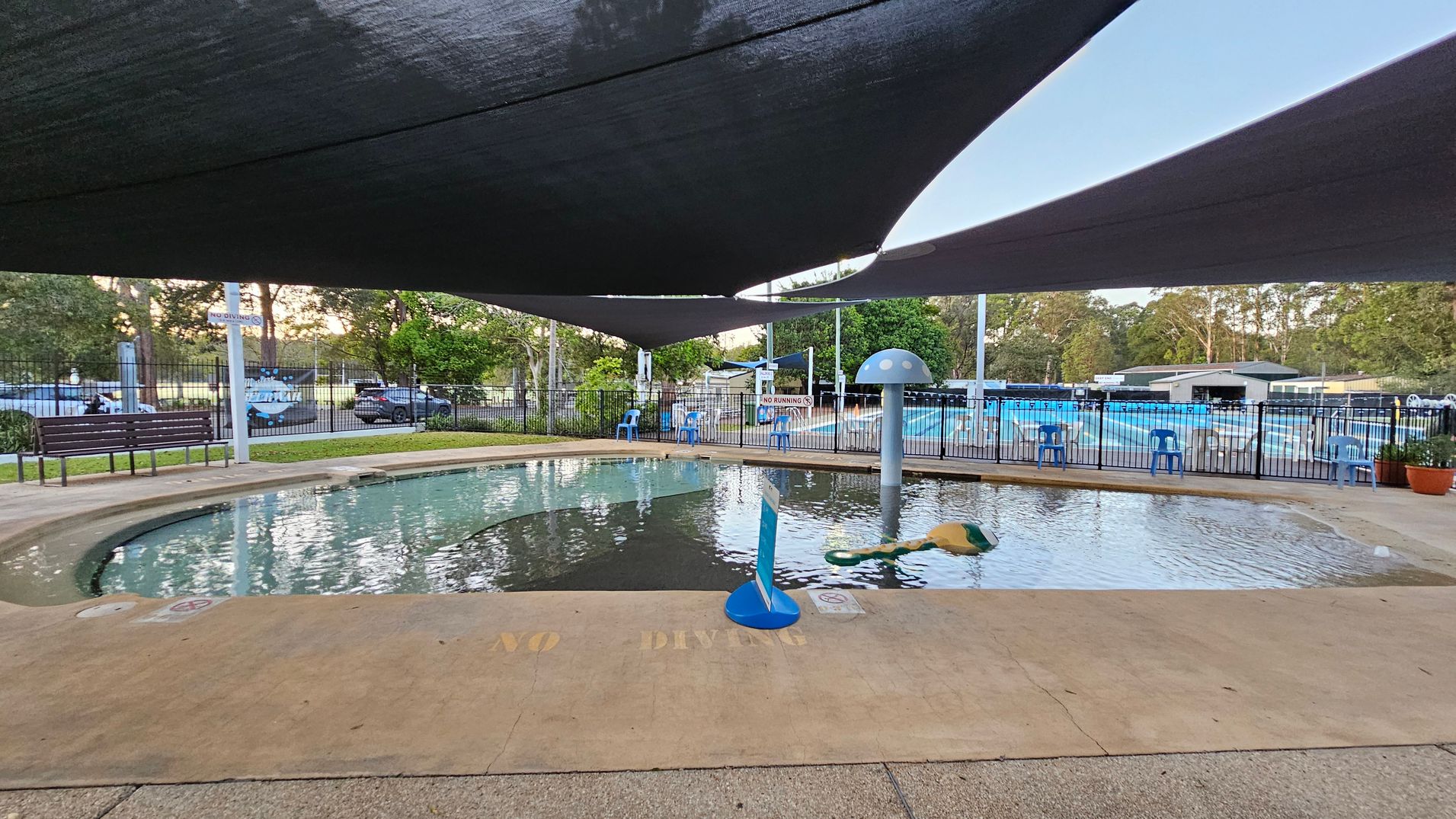 A small, shallow swimming pool is in the foreground, with a "NO DIVING" warning on the edge. A mushroom-shaped water feature is in the pool. Blue plastic chairs line the fence in the background, with a larger pool visible beyond. Shade sails cover the area above, and trees and parked cars are visible outside the fenced area. A bench is on the left side near the fence.