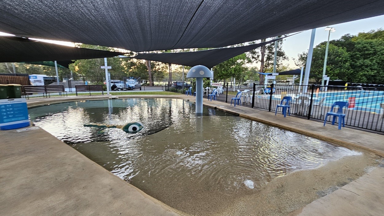 A shallow children's pool with a playful water feature shaped like a creature with eyes is in the foreground. The pool is surrounded by a concrete path. Blue plastic chairs are lined up along a black metal fence on the right. In the background, there is a larger swimming pool and some trees. Overhead, a large shade cloth provides cover. Benches and a sign are visible on the left side.