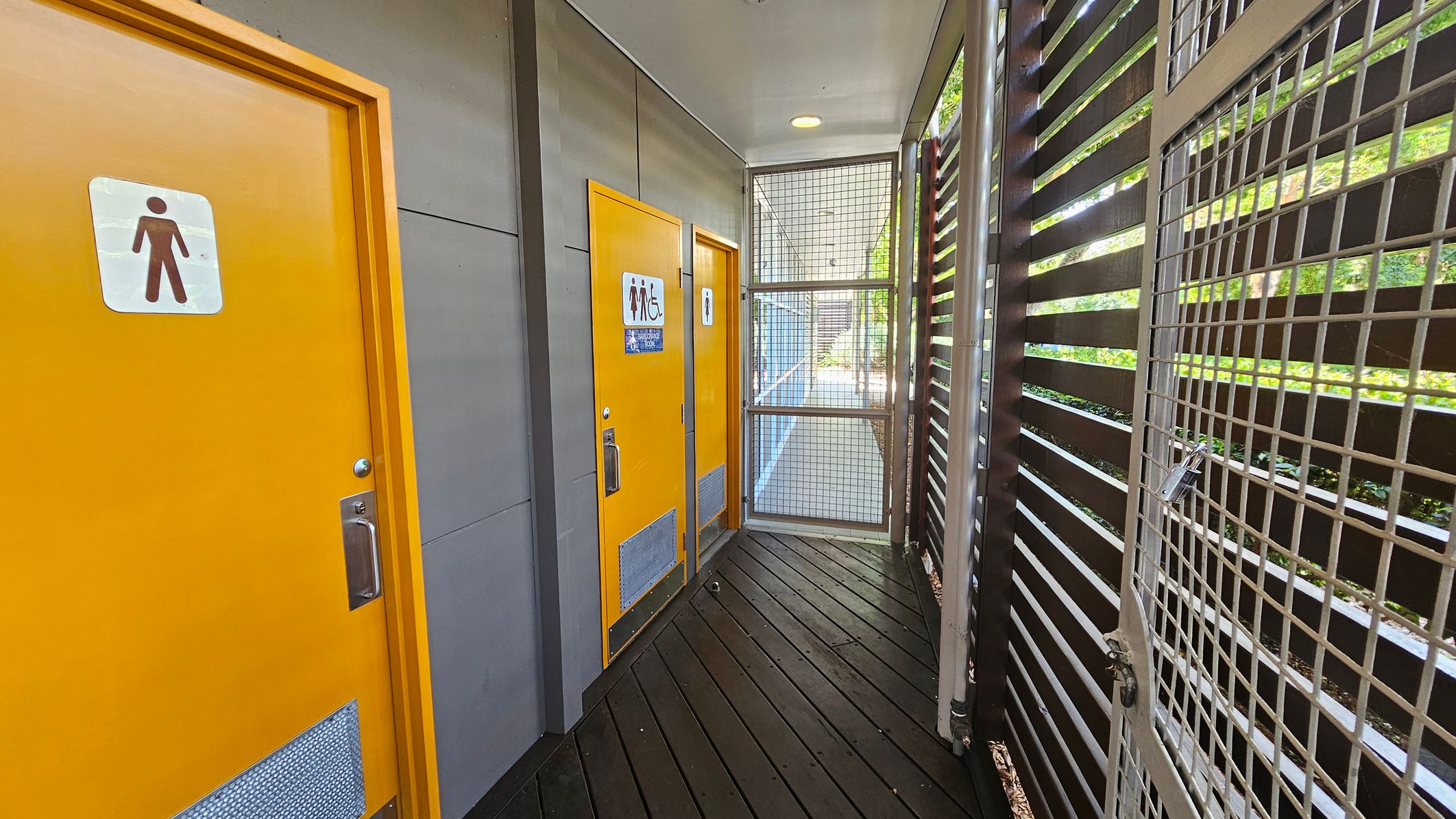 Glass entrance doors to Beerwah Library with a sign above reading "Sunshine Coast Council." Inside, there are books displayed on shelves and posters on stands. The floor is tiled, and a black rubber mat is placed outside on the wooden deck. To the right, a plaque is mounted on the wall, and a bin is visible. The left side shows a glimpse of greenery through a window.