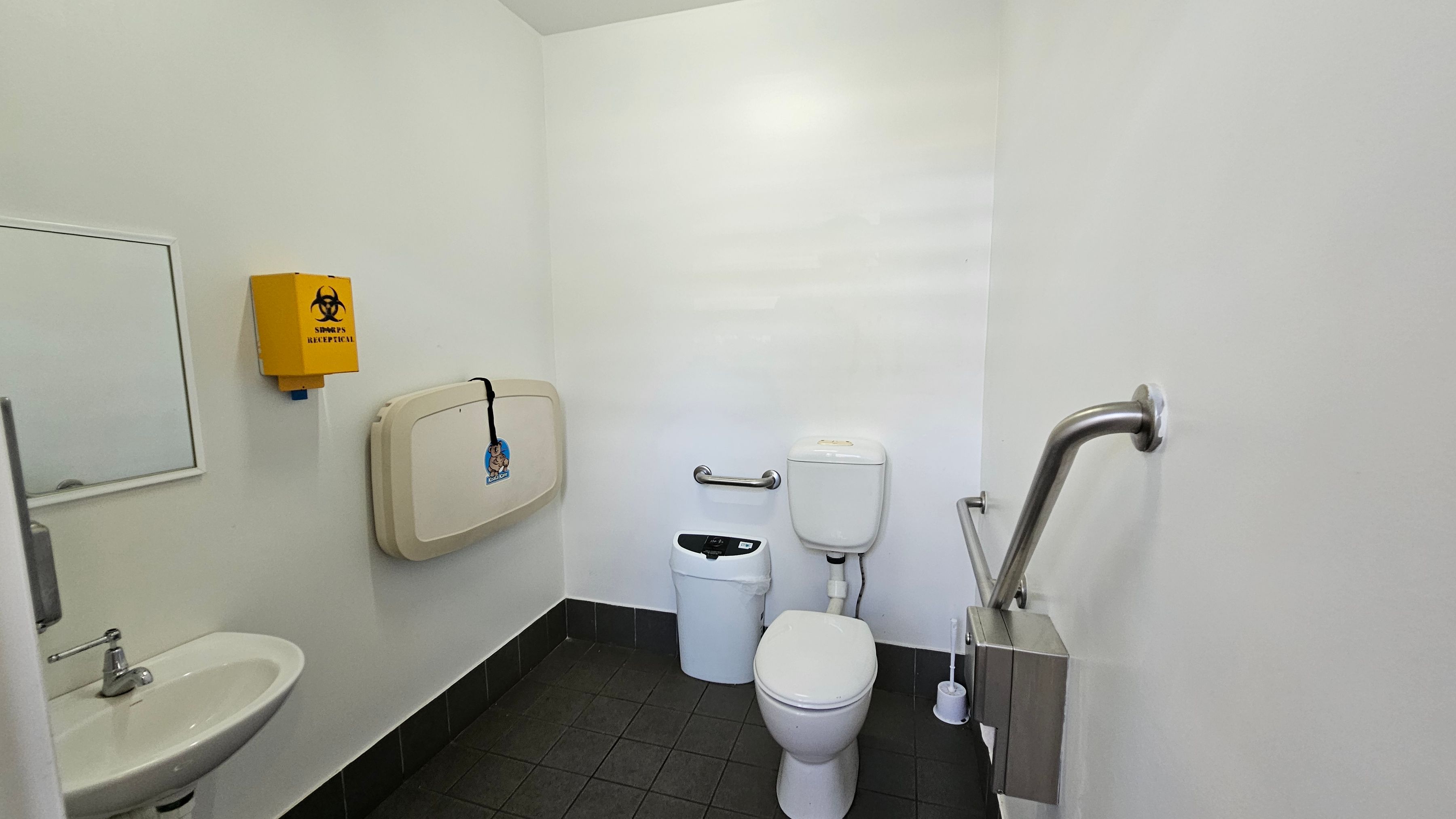 A public restroom with white walls and a dark tiled floor. On the left, there's a small sink with a mirror above it and a soap dispenser. A yellow biohazard disposal box is mounted on the wall. A baby changing station is also attached to the wall. In the centre, there's a toilet with a grab rail on the right side and a small bin nearby. A toilet brush is placed on the floor next to the toilet. The ceiling is plain and light-coloured.