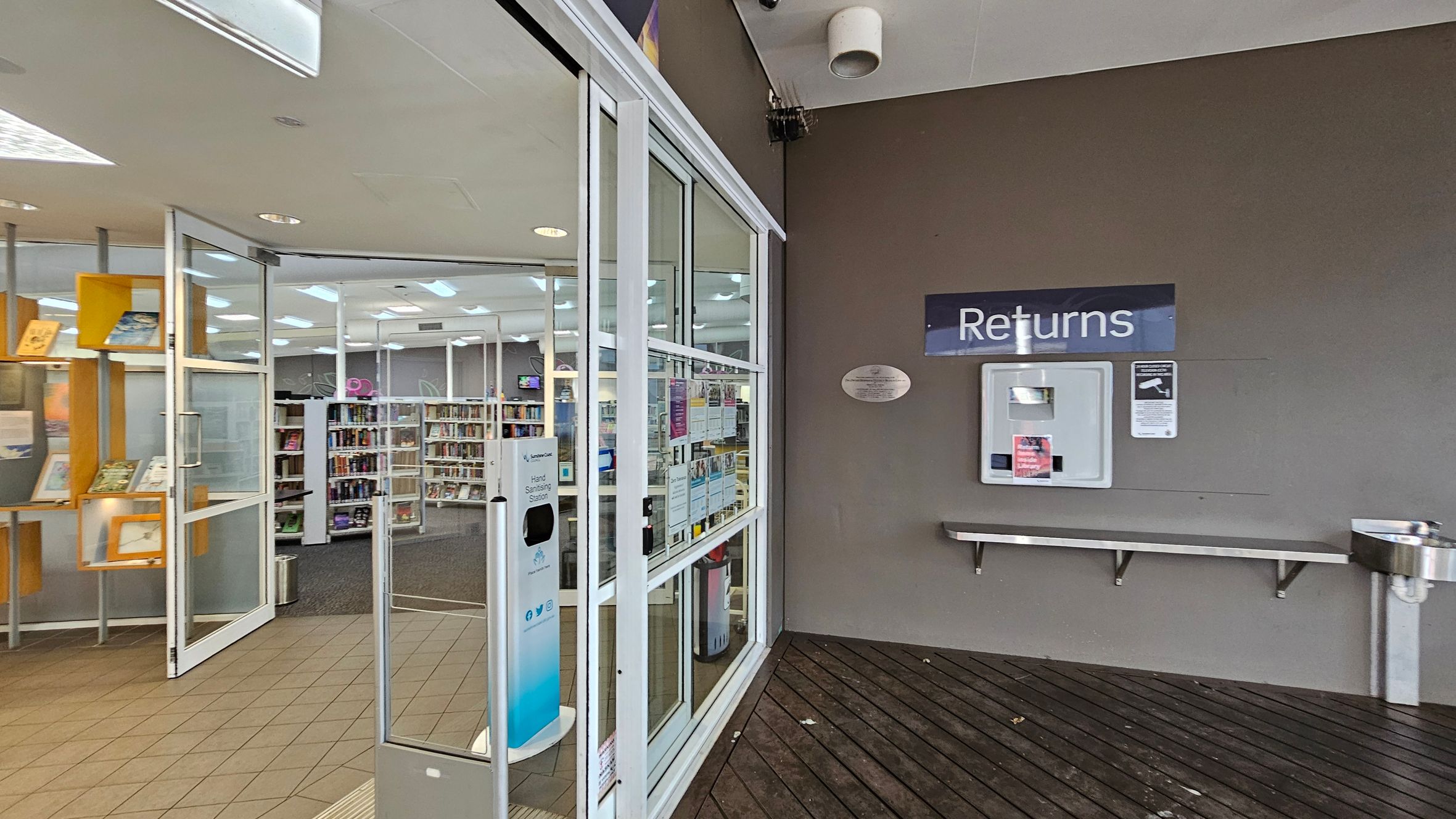 On the left, there is a library entrance with glass doors, displaying books on wooden shelves inside. A hand sanitising station is positioned near the entrance. On the right, a wall features a "Returns" slot for book returns, with a small shelf and a drinking fountain below. The floor is a combination of tiles near the entrance and wooden planks near the returns area. The ceiling is light-coloured with recessed lighting.