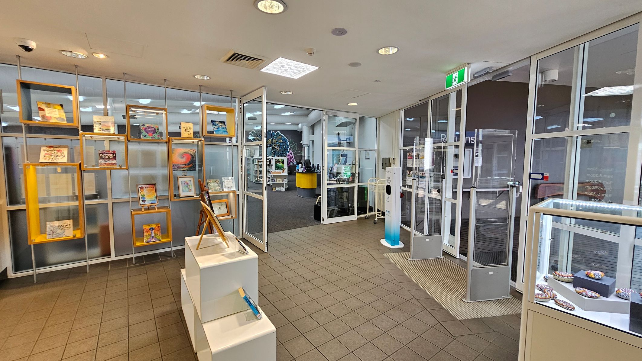 A library entrance with a tiled floor and a ceiling with recessed lights. On the left, there are display shelves with books and artwork in wooden frames. In the centre, glass doors lead into the library, where a yellow circular desk and bookshelves are visible. On the right, a glass display case contains decorative items. An exit sign is above the doors, and a hand sanitiser station is near the entrance.
