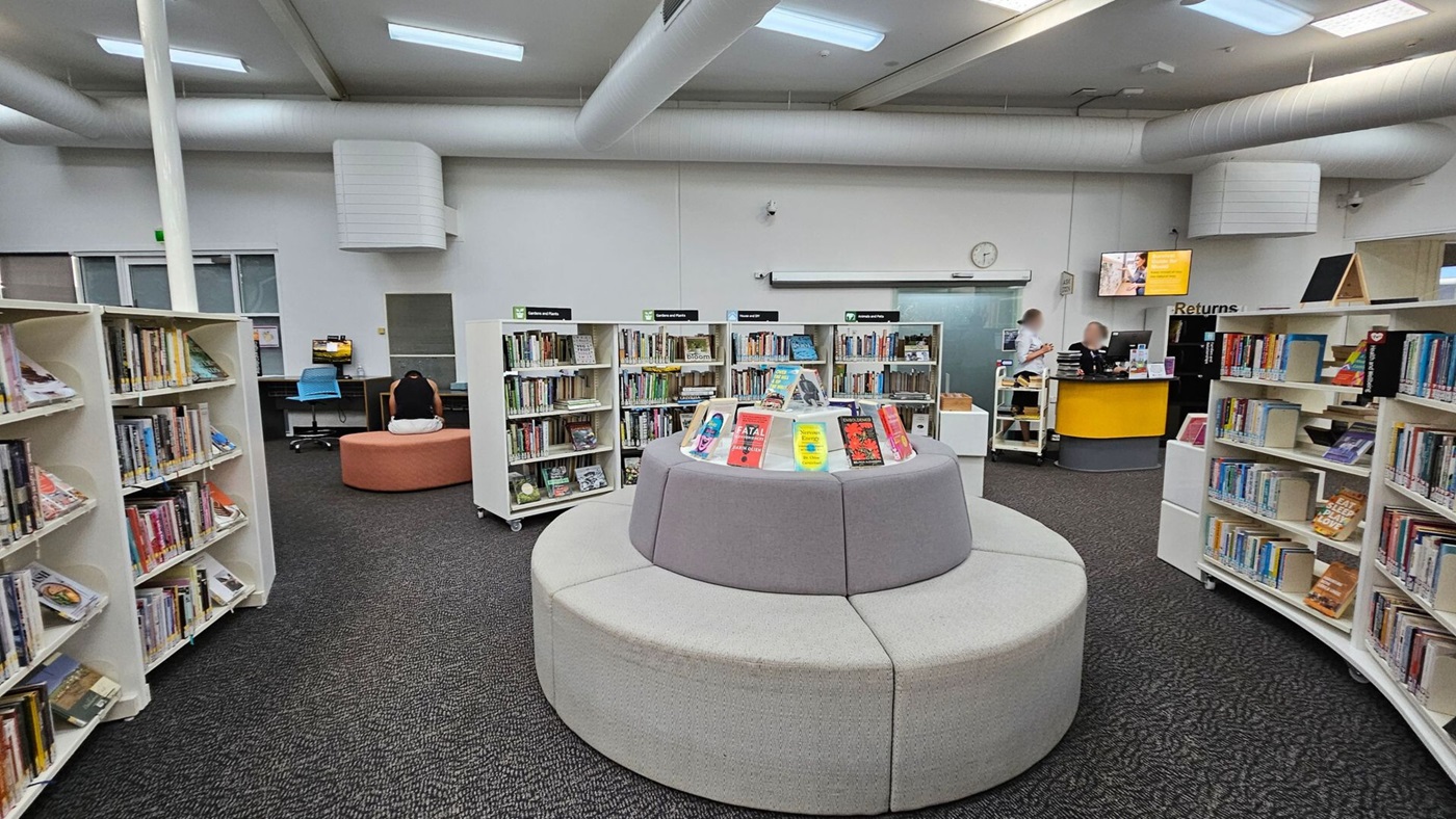 A library interior with grey carpeted flooring and a high ceiling with exposed ducts. In the centre, a circular grey seating area surrounds a display of books. Bookshelves filled with books line the left and right sides. In the background, a person sits on a round orange seat near a computer desk with a blue chair. To the right, two people are at a yellow and black returns counter, with a digital screen above them.