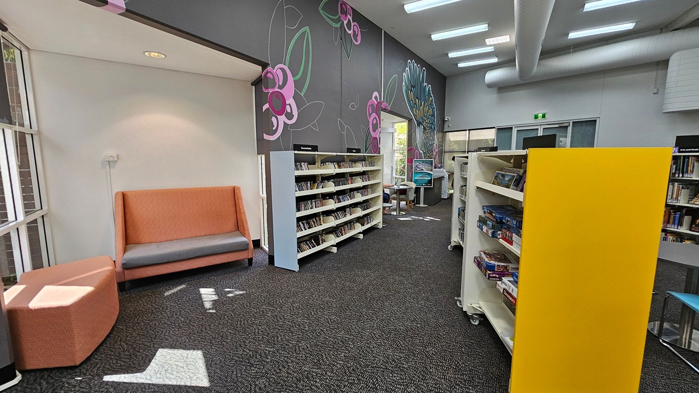 A library interior with a grey carpeted floor. On the left, there's an orange cushioned bench with a grey seat and a matching ottoman. The wall behind has large floral designs in pink and green. In the centre, there are white bookshelves filled with books and a yellow end panel. In the background, more shelves and a digital screen are visible. The ceiling has white panels and large air ducts. Natural light enters through windows on the left.