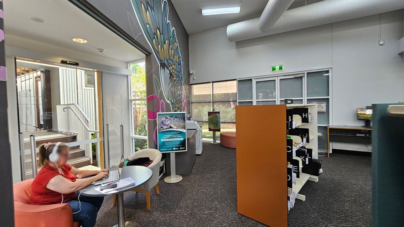 A room with a person in a red shirt and headphones sitting at a small round table on the left, using a laptop. Behind them is a glass door with a view of stairs outside. A large mural of a bird decorates the wall. In the centre, there's a sign on a stand. On the right, a bookshelf with labelled sections and a computer on a desk. The floor is carpeted, and the ceiling has exposed ducts and lighting.