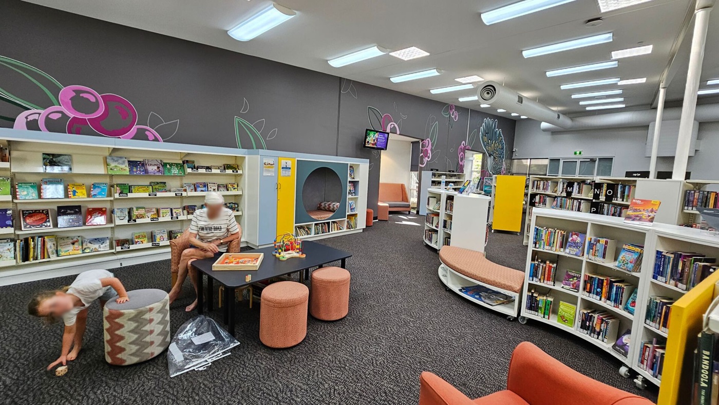 A library interior with a dark carpeted floor and a ceiling with fluorescent lights. On the left, a child is playing near a patterned ottoman, while an adult sits at a table with games. Behind them are bookshelves filled with children's books. The back wall features a mural with pink and green floral designs. On the right, more curved bookshelves hold various books, and a yellow partition is visible. A small seating area is in the background.