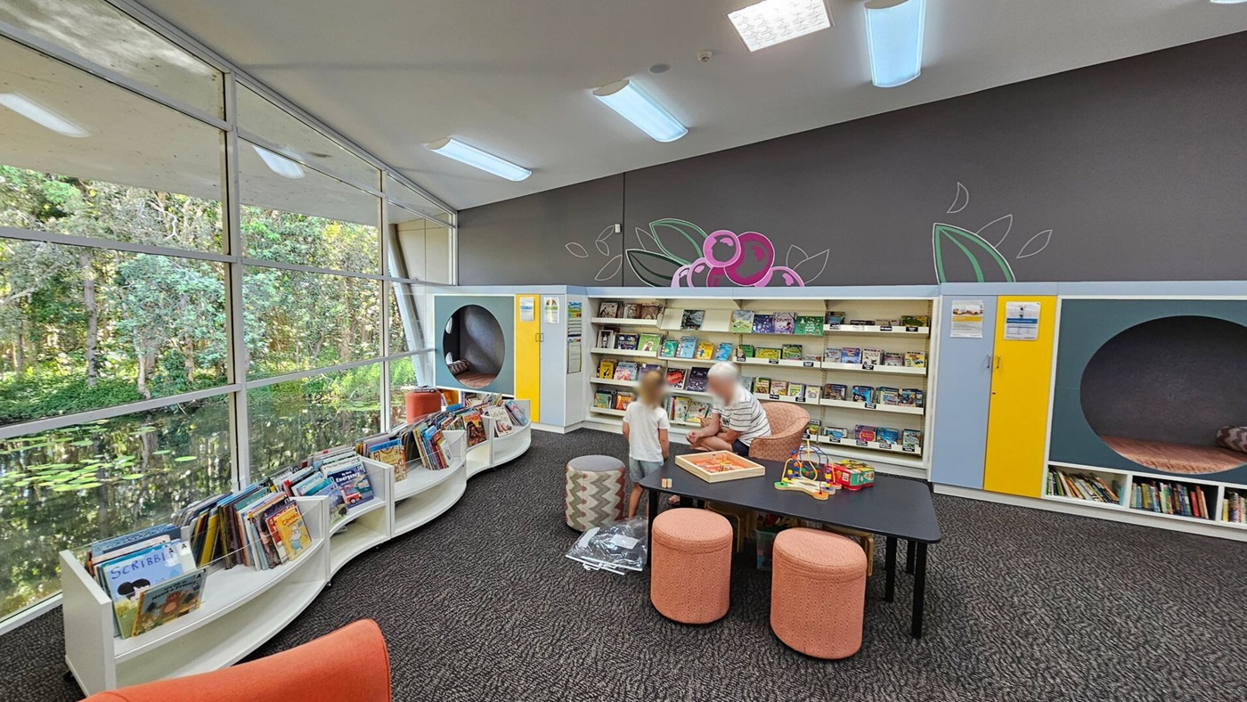 A library with large windows on the left, offering a view of a lush, green forest and a pond. Inside, there are white, curved bookshelves filled with children's books. In the centre, a child and an adult are engaged in an activity at a black table surrounded by orange stools. The back wall features bookshelves, a colourful mural, and circular reading nooks. The ceiling has modern light fixtures, and the floor is carpeted in dark grey.