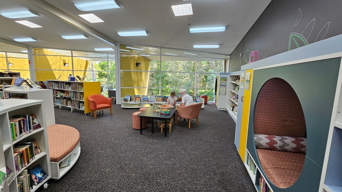 A library interior with a grey carpeted floor and a ceiling with rectangular lights. On the left, there are bookshelves filled with books and a curved seating area. In the centre, a child and an adult sit at a table with toys and books, surrounded by orange chairs. The right side features a circular reading nook with patterned cushions. Large windows in the background reveal a view of trees and a yellow wall outside.