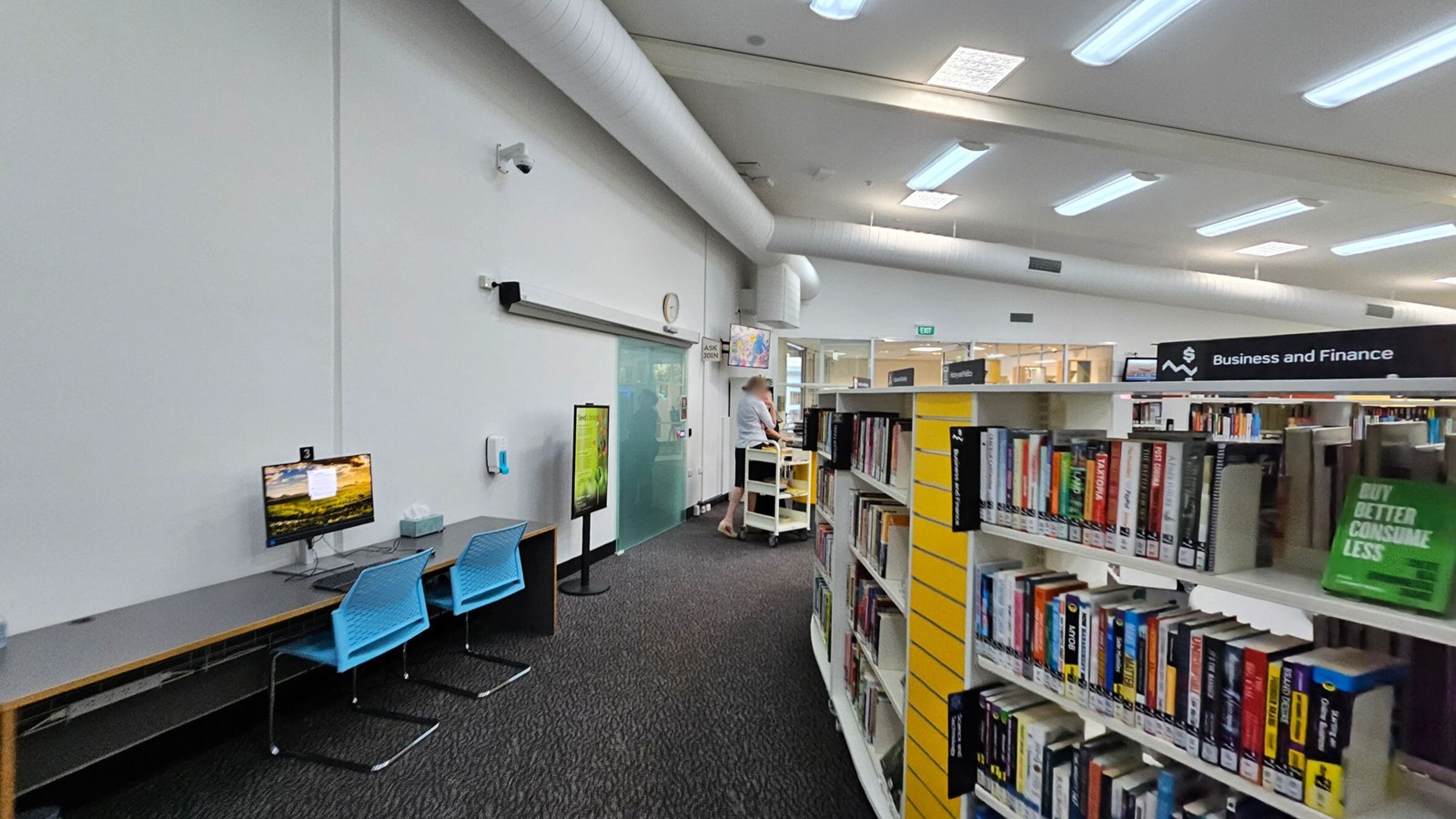 On the left, there are two blue chairs in front of a long desk with a computer monitor displaying a landscape image. A wall-mounted clock and a security camera are above. In the middle, a person stands near a service desk with a trolley. On the right, bookshelves labelled "Business and Finance" are filled with books. The ceiling has fluorescent lights and visible air ducts, and the floor is carpeted in dark grey.