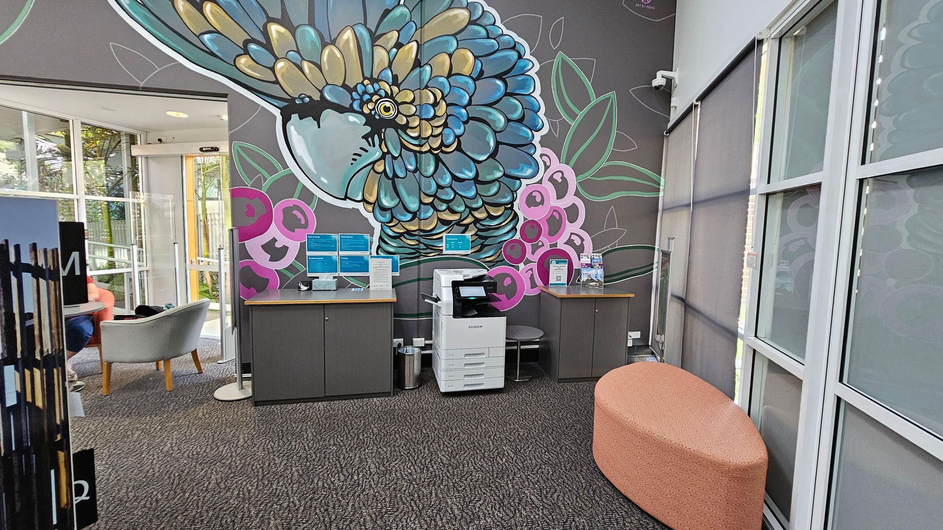 A room with a large, colourful mural of a bird on the wall. In the centre, there's a white printer on a grey cabinet, flanked by two more cabinets with informational displays. To the right, a peach-coloured, oval-shaped ottoman sits on a patterned carpet. On the left, a glass door leads to an area with grey chairs and a small table. The ceiling is not visible, and the windows have grey blinds.