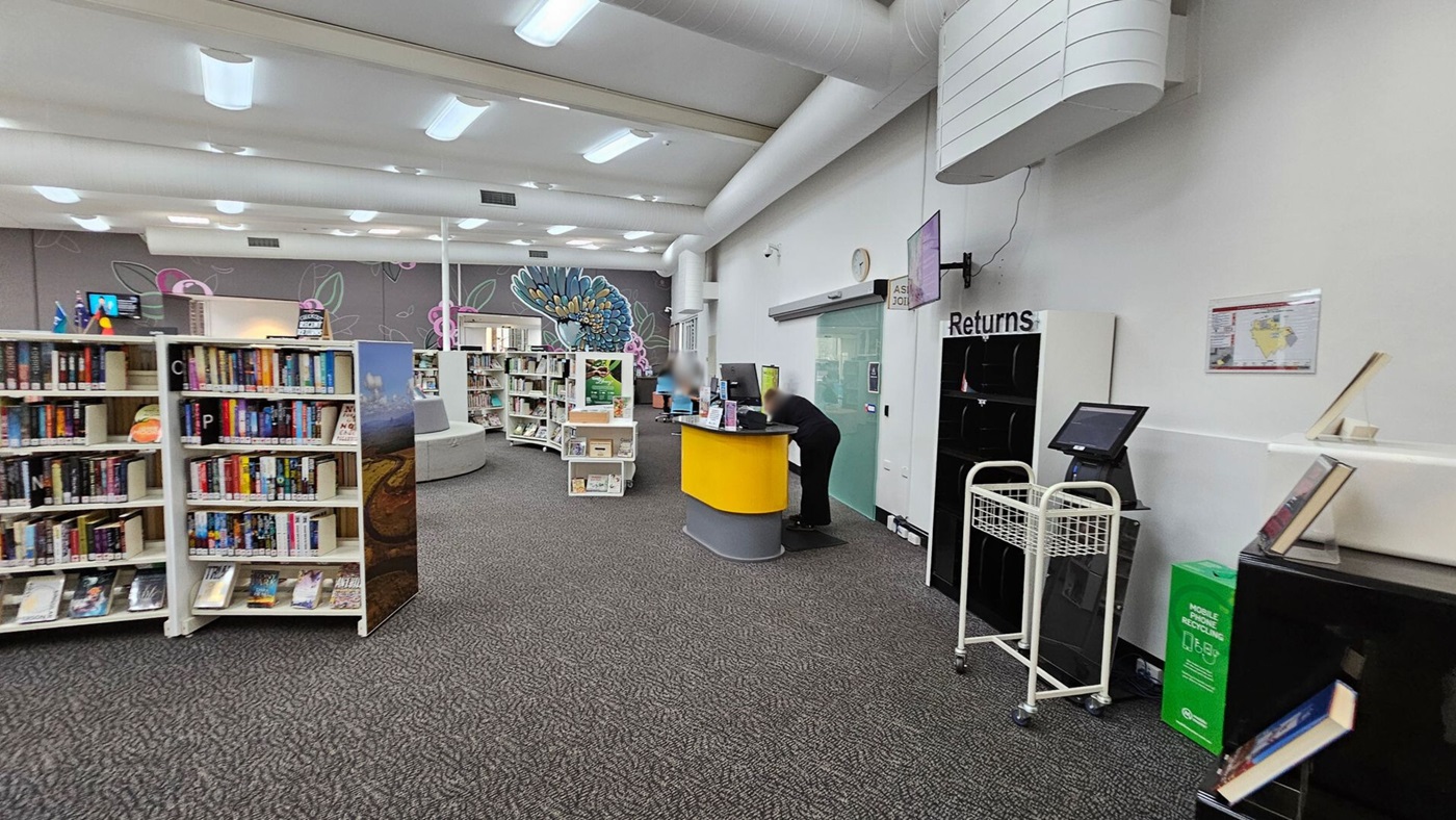 A library interior with a grey carpeted floor. On the left, there are bookshelves filled with books. In the middle, a yellow and grey desk is attended by a person. To the right, a book return area with a cart and a digital kiosk is visible. The back wall features a colourful mural with floral designs. The ceiling has white panels with fluorescent lights.