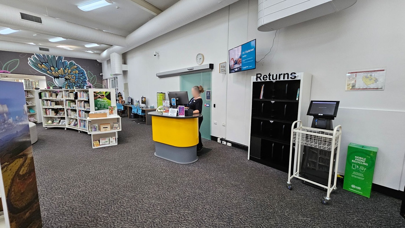 A library interior with a grey carpeted floor. On the right, a returns section with black shelves and a computer on a stand. A green mobile phone recycling bin is nearby. In the centre, a staff member stands at a yellow and grey desk with a computer. On the left, white bookshelves filled with books and a mural of a colourful bird on the wall. Blue chairs are visible near computers along the wall. The ceiling has large white ducts and fluorescent lights.