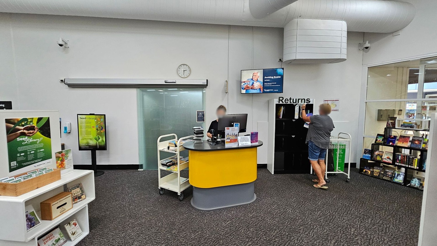 A library interior with a grey carpeted floor. On the left, there is a display with seed library information and a wooden box on a white shelf. In the centre, a person sits at a yellow and grey desk with a computer and a trolley of books. Above, a clock and a TV screen are mounted on the wall. On the right, a person is returning books at a "Returns" section. Nearby, a glass wall reveals more bookshelves and a green recycling bin. The ceiling has large white ducts.