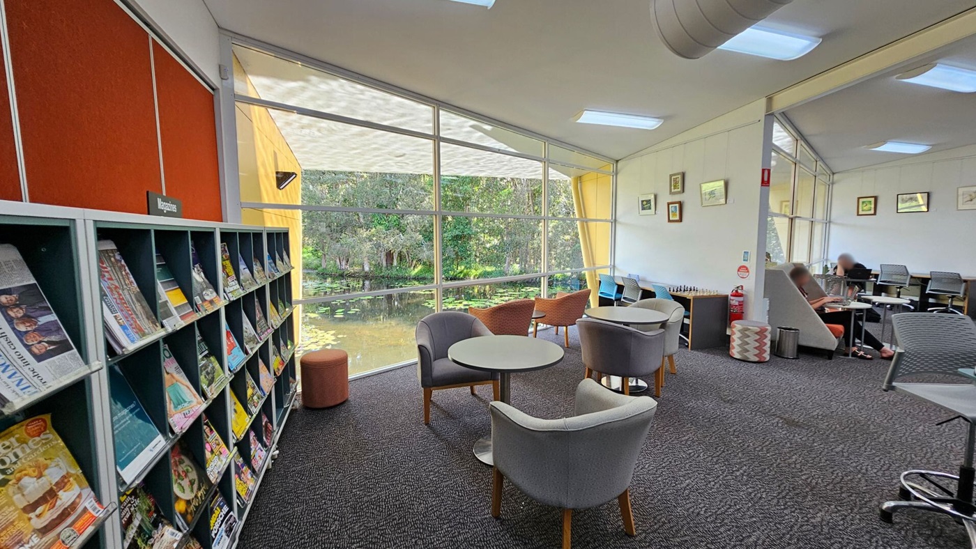 A library interior with bookshelves on the right, labelled "A" and "B", filled with books. On the left, there are shelves with magazines and a payment terminal. In the middle, a seating area with grey and brown chairs around small tables. Large windows in the background reveal a view of trees and a pond. The carpet is dark grey, and the ceiling has fluorescent lights and a visible air duct. A fire extinguisher and some framed pictures are on the wall.