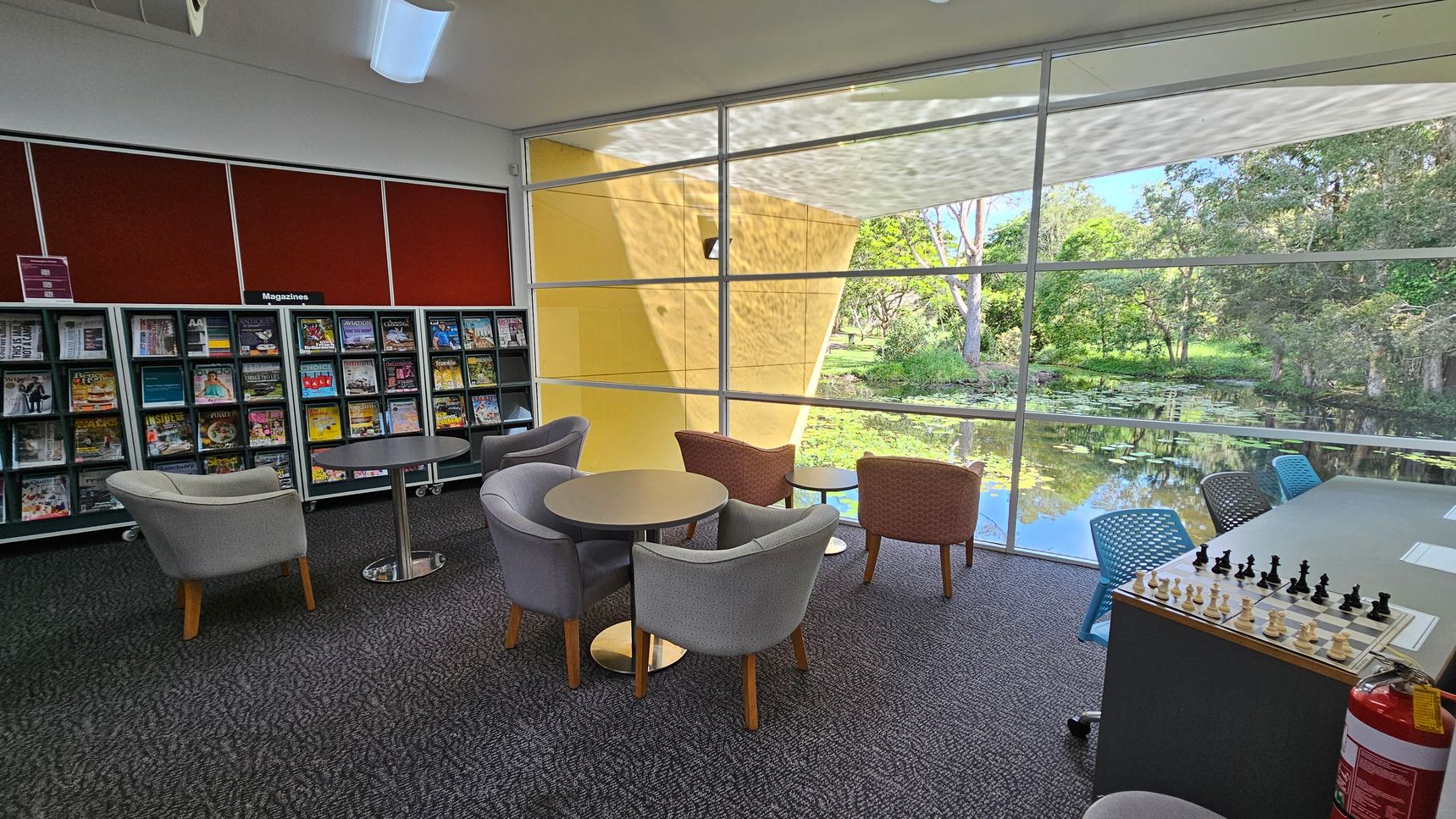 A library reading area with a carpeted floor. On the left, there are shelves filled with magazines under a sign labelled "Magazines". In the centre, several round tables are surrounded by grey and brown upholstered chairs. On the right, a chessboard is set up on a table with blue chairs. Large windows in the background offer a view of a pond with lily pads and trees, allowing natural light to fill the space.