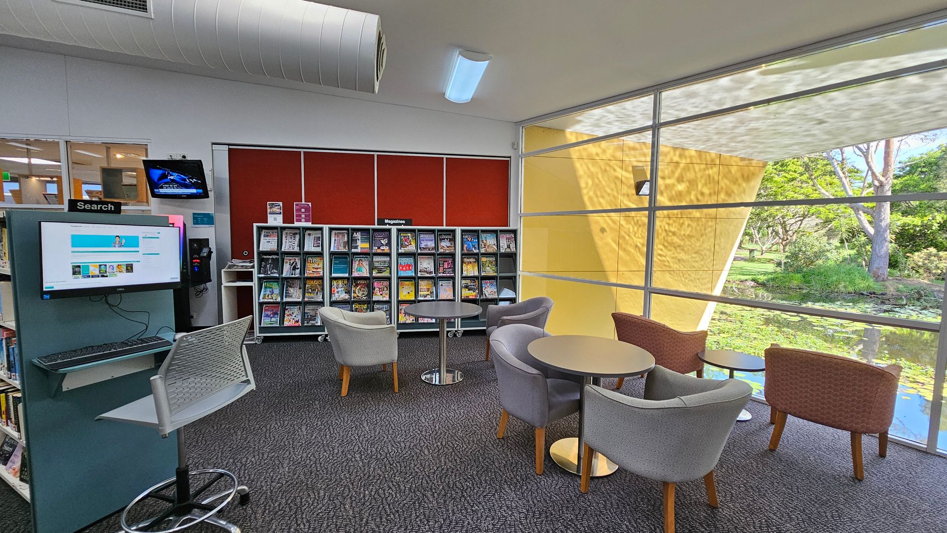 A library interior with a search computer station on the left, featuring a monitor and a chair. Behind it, a wall-mounted TV is visible. In the centre, there is a magazine rack filled with various titles against a red wall. On the right, there are round tables with grey and brown chairs. Large windows provide a view of a garden with trees and a pond outside. The floor is carpeted, and the ceiling has a cylindrical air duct and lighting fixtures.