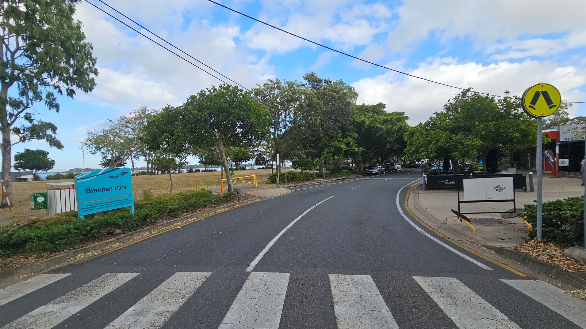A road curves to the right with a zebra crossing in the foreground. On the left, a sign reads "Brennan Park" next to a grassy area with trees. In the background, there are more trees and a glimpse of water. On the right, a pedestrian crossing sign is visible, along with a footpath and a building with signage. The area is lined with trees and shrubs, and there are parked cars further down the road. A road curves to the right with a zebra crossing in the foreground. On the left, a sign reads "Brennan Park" next to a grassy area with trees. In the background, there are more trees and a glimpse of water. On the right, a pedestrian crossing sign is visible, along with a footpath and a building with signage. The area is lined with trees and shrubs, and there are parked cars further down the road.
