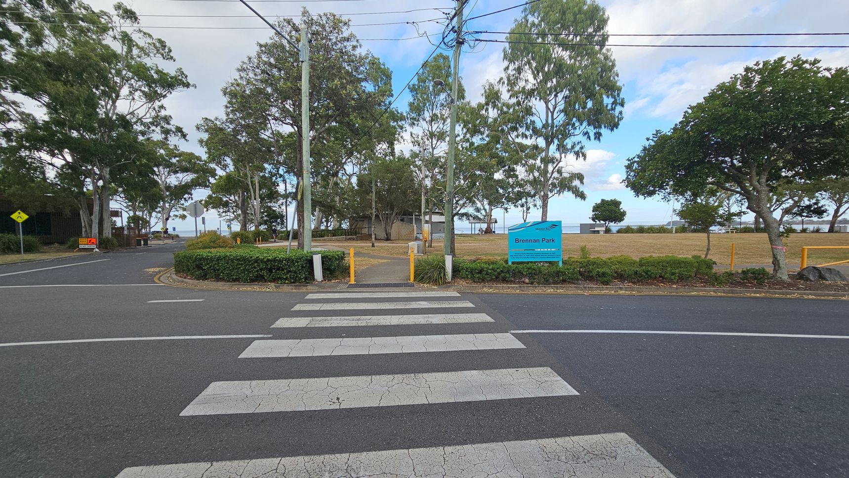 A zebra crossing leads to a park entrance with a blue sign reading "Brennan Park" on the right. The park is bordered by trees and has a grassy area. Yellow bollards and a path are visible beyond the crossing. Power lines run overhead, and there are more trees and a road to the left. The background shows a glimpse of water beyond the park. A zebra crossing leads to a park entrance with a blue sign reading "Brennan Park" on the right. The park is bordered by trees and has a grassy area. Yellow bollards and a path are visible beyond the crossing. Power lines run overhead, and there are more trees and a road to the left. The background shows a glimpse of water beyond the park.