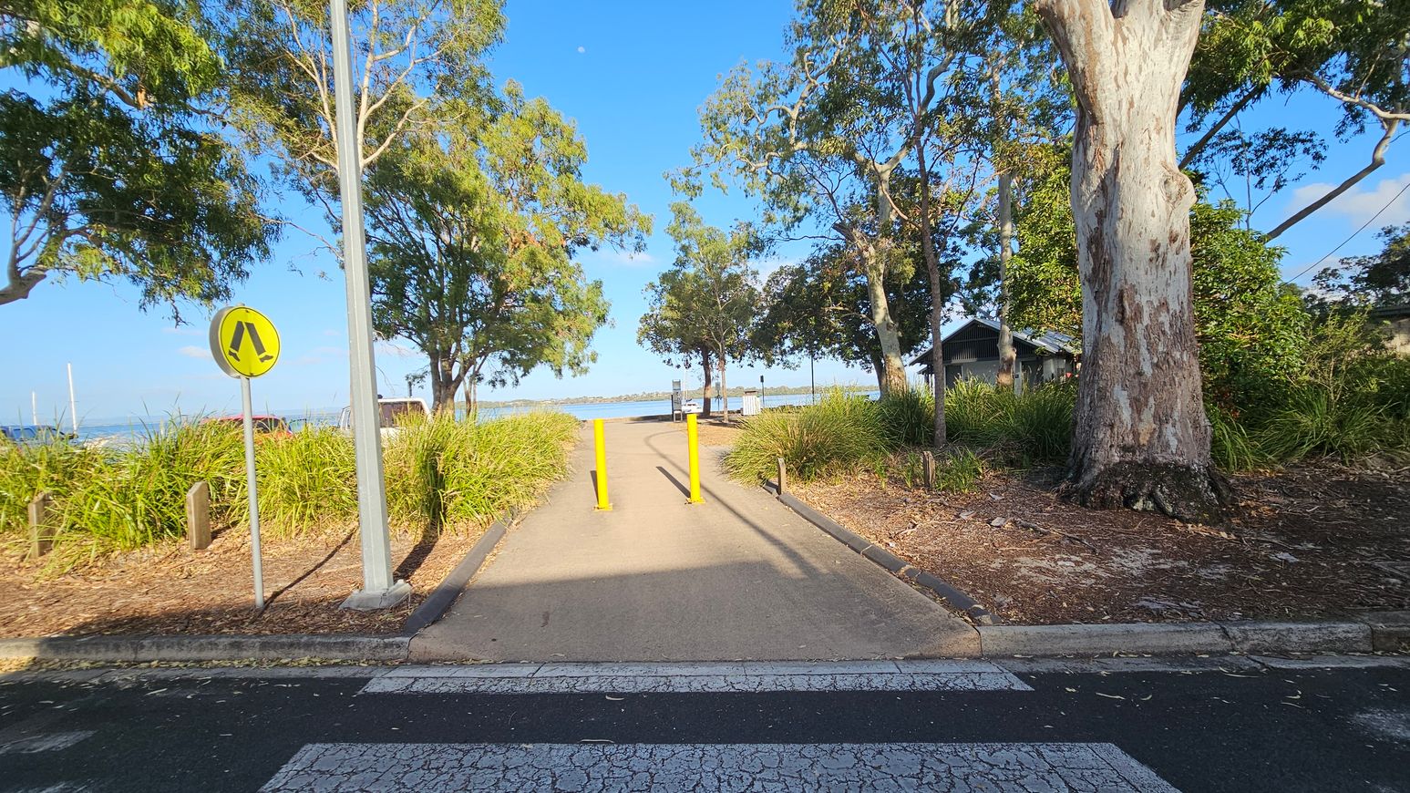 A pathway leads towards a waterfront, flanked by tall trees and dense grass on both sides. A pedestrian crossing sign is on the left, and a large tree trunk is on the right. Yellow bollards are positioned in the middle of the path. In the background, a body of water is visible, with a small structure to the right. The ground is paved, and the area is shaded by the trees. A pathway leads towards a waterfront, flanked by tall trees and dense grass on both sides. A pedestrian crossing sign is on the left, and a large tree trunk is on the right. Yellow bollards are positioned in the middle of the path. In the background, a body of water is visible, with a small structure to the right. The ground is paved, and the area is shaded by the trees.