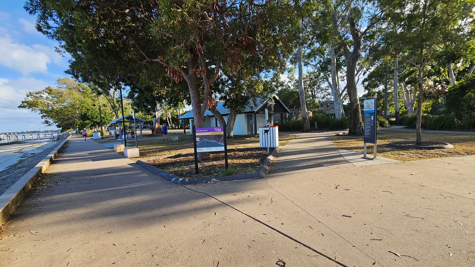 A paved walkway runs along the left side, bordered by a low stone wall and a body of water. Trees line the path, providing shade. In the centre, a signpost and a small building with a sloped roof are visible. On the right, another signpost stands near a grassy area with more trees. A person walks a dog in the distance. The ground is covered with scattered leaves and the area is well-lit by natural light. A paved walkway runs along the left side, bordered by a low stone wall and a body of water. Trees line the path, providing shade. In the centre, a signpost and a small building with a sloped roof are visible. On the right, another signpost stands near a grassy area with more trees. A person walks a dog in the distance. The ground is covered with scattered leaves and the area is well-lit by natural light.