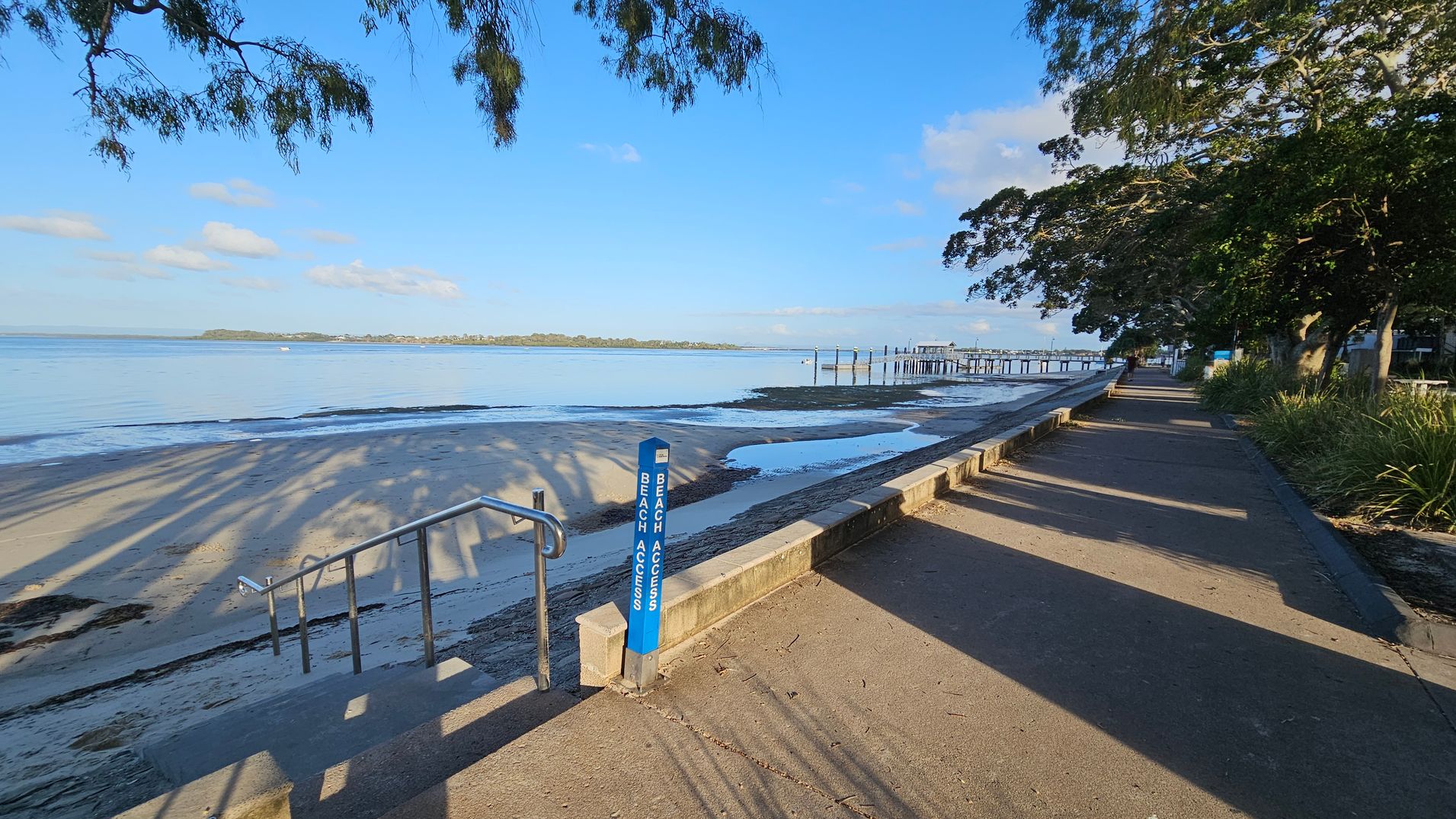 A coastal scene with a sandy beach on the left, leading to calm blue water. A metal railing provides access to the beach, marked by a blue "Beach Access" sign. On the right, a paved walkway is lined with trees casting long shadows. In the distance, a pier extends into the water. The background features a clear sky with a few clouds and a distant shoreline. A coastal scene with a sandy beach on the left, leading to calm blue water. A metal railing provides access to the beach, marked by a blue "Beach Access" sign. On the right, a paved walkway is lined with trees casting long shadows. In the distance, a pier extends into the water. The background features a clear sky with a few clouds and a distant shoreline.