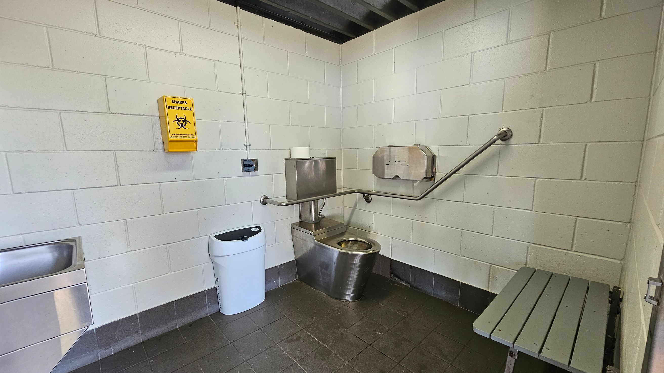 A public restroom with white tiled walls and a dark tiled floor. On the left, a stainless steel sink is mounted on the wall. In the centre, a stainless steel toilet with a roll of toilet paper on top is accompanied by a wall-mounted paper dispenser and a grab bar. A yellow sharps receptacle is fixed to the wall above. A white bin is positioned near the toilet. On the right, a grey bench is attached to the wall. The ceiling is dark with visible beams. A public restroom with white tiled walls and a dark tiled floor. On the left, a stainless steel sink is mounted on the wall. In the centre, a stainless steel toilet with a roll of toilet paper on top is accompanied by a wall-mounted paper dispenser and a grab bar. A yellow sharps receptacle is fixed to the wall above. A white bin is positioned near the toilet. On the right, a grey bench is attached to the wall. The ceiling is dark with visible beams.