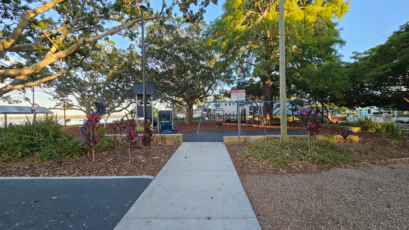 A playground area with a concrete path leading to swings and play equipment. On the left, there are trees with thick branches and a glimpse of a waterfront with a shelter. On the right, more trees and a parking area are visible. The ground is covered with wood chips and small plants, and there are two tall poles near the path. The background features a mix of greenery and buildings. A playground area with a concrete path leading to swings and play equipment. On the left, there are trees with thick branches and a glimpse of a waterfront with a shelter. On the right, more trees and a parking area are visible. The ground is covered with wood chips and small plants, and there are two tall poles near the path. The background features a mix of greenery and buildings.