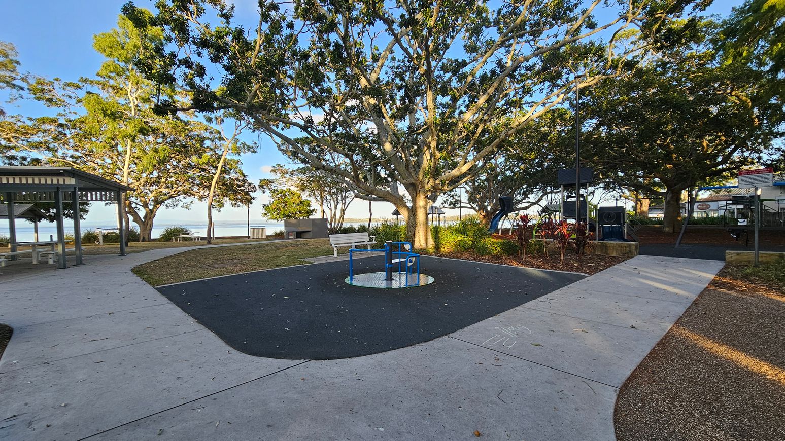 A park scene with a large tree in the centre, surrounded by a paved path. In the middle, there's a blue and white roundabout on a dark rubber surface. To the left, a sheltered picnic area with tables and benches. On the right, a playground with climbing equipment and a slide. The background features more trees and a glimpse of water. The ground is a mix of concrete, grass, and mulch, with scattered leaves. A park scene with a large tree in the centre, surrounded by a paved path. In the middle, there's a blue and white roundabout on a dark rubber surface. To the left, a sheltered picnic area with tables and benches. On the right, a playground with climbing equipment and a slide. The background features more trees and a glimpse of water. The ground is a mix of concrete, grass, and mulch, with scattered leaves.