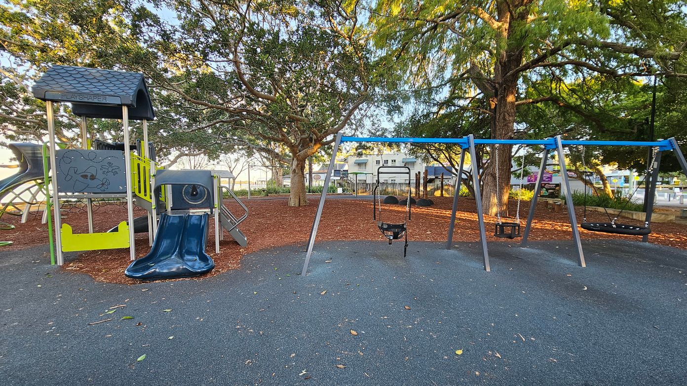 A playground with a play structure on the left featuring a small roof, climbing elements, and a blue slide. To the right, there is a swing set with multiple swings. The ground is covered with a dark rubber surface, and the area is surrounded by trees with dense foliage. In the background, there are buildings and a fence, with some benches and signs visible. The scene is well-lit, suggesting daylight. A playground with a play structure on the left featuring a small roof, climbing elements, and a blue slide. To the right, there is a swing set with multiple swings. The ground is covered with a dark rubber surface, and the area is surrounded by trees with dense foliage. In the background, there are buildings and a fence, with some benches and signs visible. The scene is well-lit, suggesting daylight.