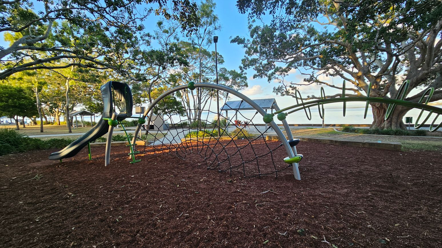 A playground with a slide and climbing net structure on the left, set on a surface covered with reddish-brown mulch. In the background, there are large trees with sprawling branches and a small pavilion with a metal roof. Beyond the trees, a body of water is visible. The area is surrounded by greenery and a paved path runs through the scene. A playground with a slide and climbing net structure on the left, set on a surface covered with reddish-brown mulch. In the background, there are large trees with sprawling branches and a small pavilion with a metal roof. Beyond the trees, a body of water is visible. The area is surrounded by greenery and a paved path runs through the scene.