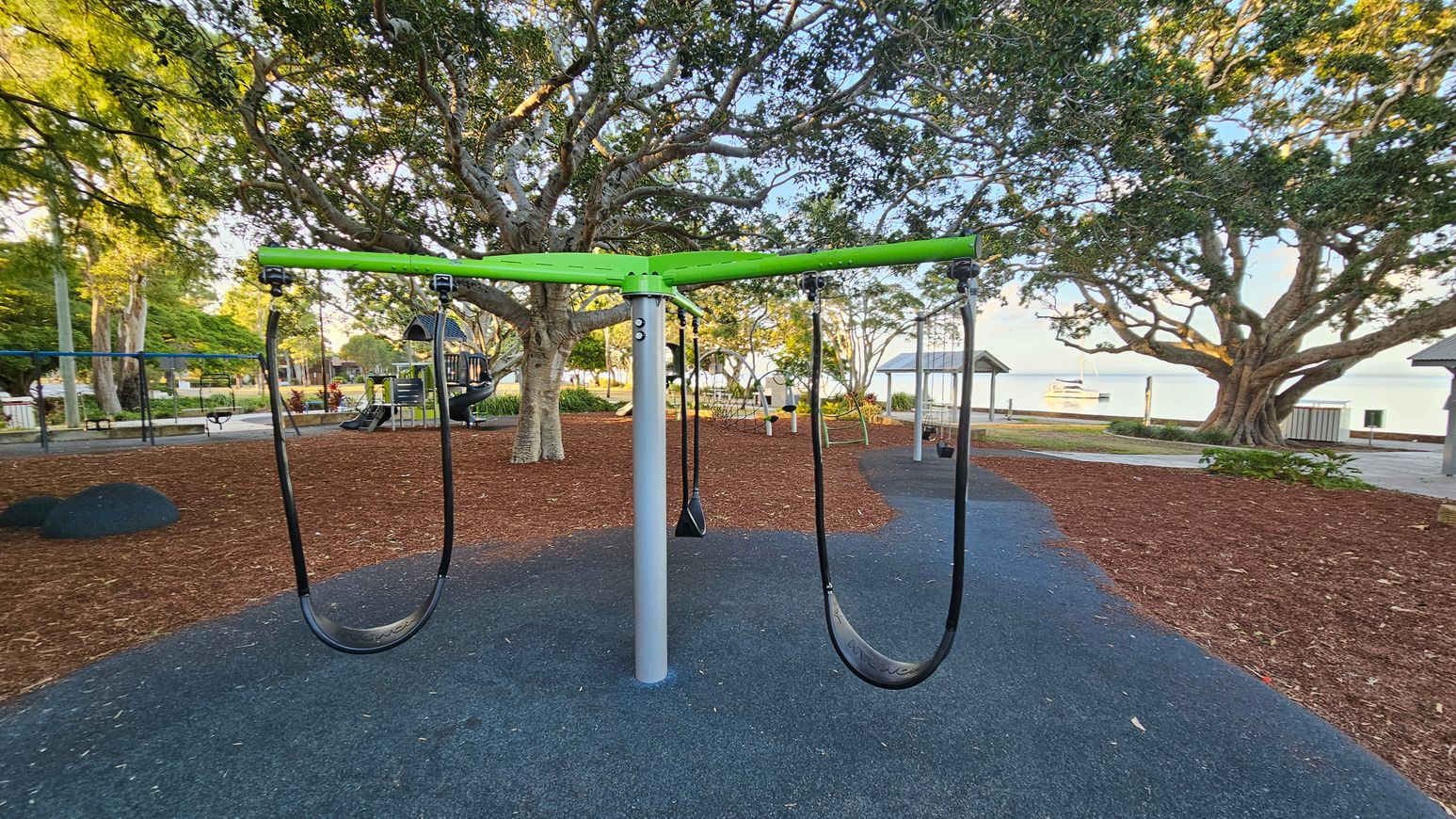 A playground with a central swing set featuring a green top and black seats. The ground is covered with a mix of dark rubber and brown mulch. In the background, large trees provide shade, and a play structure with a slide is visible. To the right, a pavilion and a glimpse of water with a boat can be seen. The area is surrounded by greenery and pathways. A playground with a central swing set featuring a green top and black seats. The ground is covered with a mix of dark rubber and brown mulch. In the background, large trees provide shade, and a play structure with a slide is visible. To the right, a pavilion and a glimpse of water with a boat can be seen. The area is surrounded by greenery and pathways.