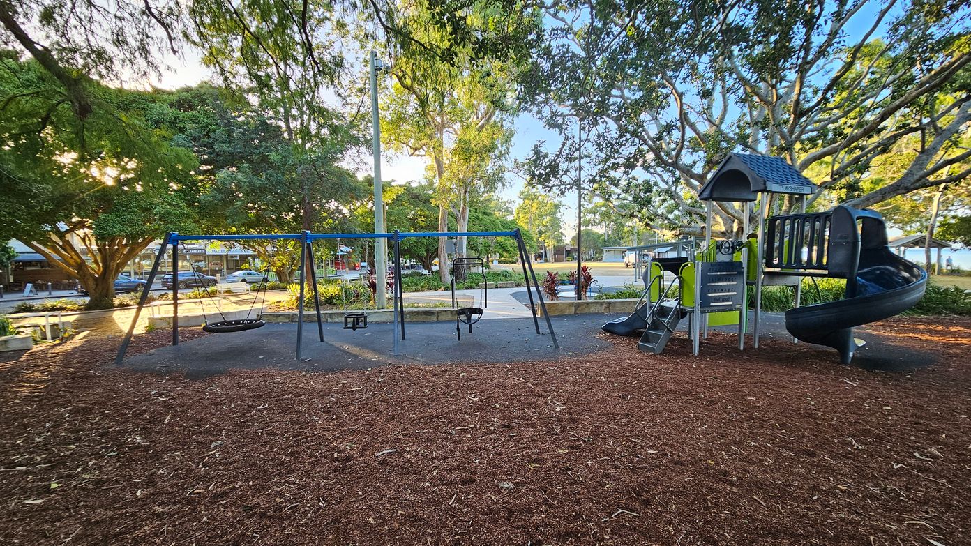 A playground with a swing set on the left, featuring a basket swing and two regular swings. On the right, there's a play structure with a slide and climbing elements. The ground is covered with wood chips, and large trees with dense foliage surround the area. In the background, there are more trees, a pathway, and a few parked cars. The sunlight filters through the leaves, creating dappled light on the ground. A playground with a swing set on the left, featuring a basket swing and two regular swings. On the right, there's a play structure with a slide and climbing elements. The ground is covered with wood chips, and large trees with dense foliage surround the area. In the background, there are more trees, a pathway, and a few parked cars. The sunlight filters through the leaves, creating dappled light on the ground.