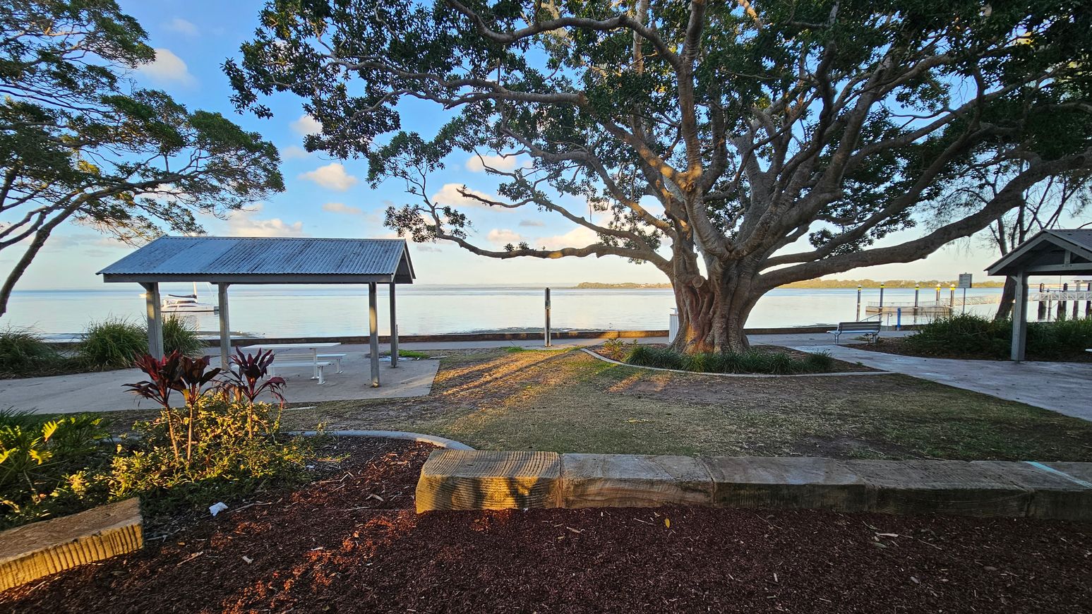A large tree with sprawling branches dominates the centre, casting shadows on the ground. To the left, a small shelter with a corrugated metal roof stands near the water's edge. On the right, another similar shelter is partially visible. The foreground features a landscaped area with red mulch and plants. The background shows a calm body of water with a boat and a distant shoreline. The ground is a mix of grass and paved paths. A large tree with sprawling branches dominates the centre, casting shadows on the ground. To the left, a small shelter with a corrugated metal roof stands near the water's edge. On the right, another similar shelter is partially visible. The foreground features a landscaped area with red mulch and plants. The background shows a calm body of water with a boat and a distant shoreline. The ground is a mix of grass and paved paths.