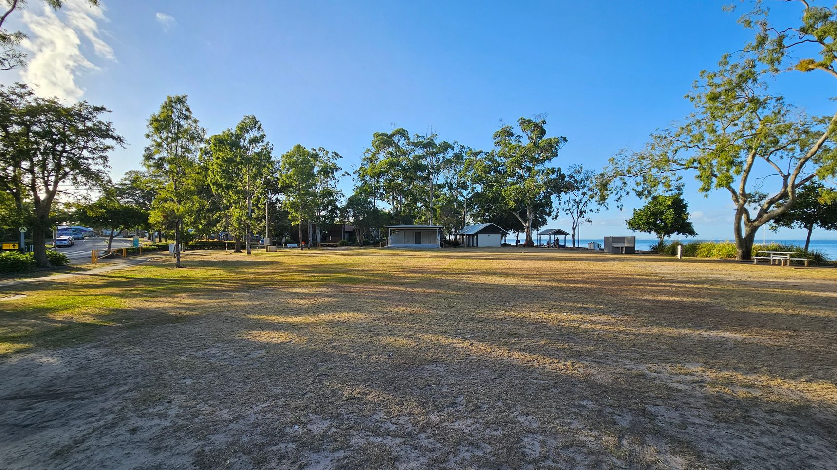 A grassy park area with patches of dry grass in the foreground. In the middle, there are several trees providing shade, and a few small structures, possibly shelters or kiosks. To the left, a road with a car and more trees. On the right, a picnic table and a view of the water in the background. The sky is clear and blue. A grassy park area with patches of dry grass in the foreground. In the middle, there are several trees providing shade, and a few small structures, possibly shelters or kiosks. To the left, a road with a car and more trees. On the right, a picnic table and a view of the water in the background. The sky is clear and blue.