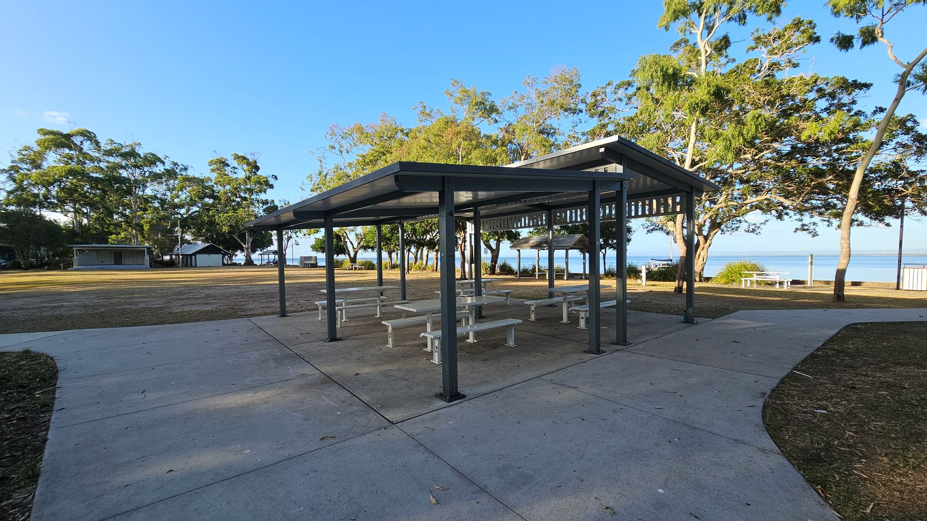 A covered picnic area with several white benches and tables is in the centre. The structure has a metal roof supported by dark-coloured poles. Surrounding the area is a concrete path with patches of grass and mulch. In the background, there are trees and a glimpse of water, suggesting a coastal location. To the left, there are small buildings, possibly restrooms or storage facilities. The area is well-maintained and open. A covered picnic area with several white benches and tables is in the centre. The structure has a metal roof supported by dark-coloured poles. Surrounding the area is a concrete path with patches of grass and mulch. In the background, there are trees and a glimpse of water, suggesting a coastal location. To the left, there are small buildings, possibly restrooms or storage facilities. The area is well-maintained and open.