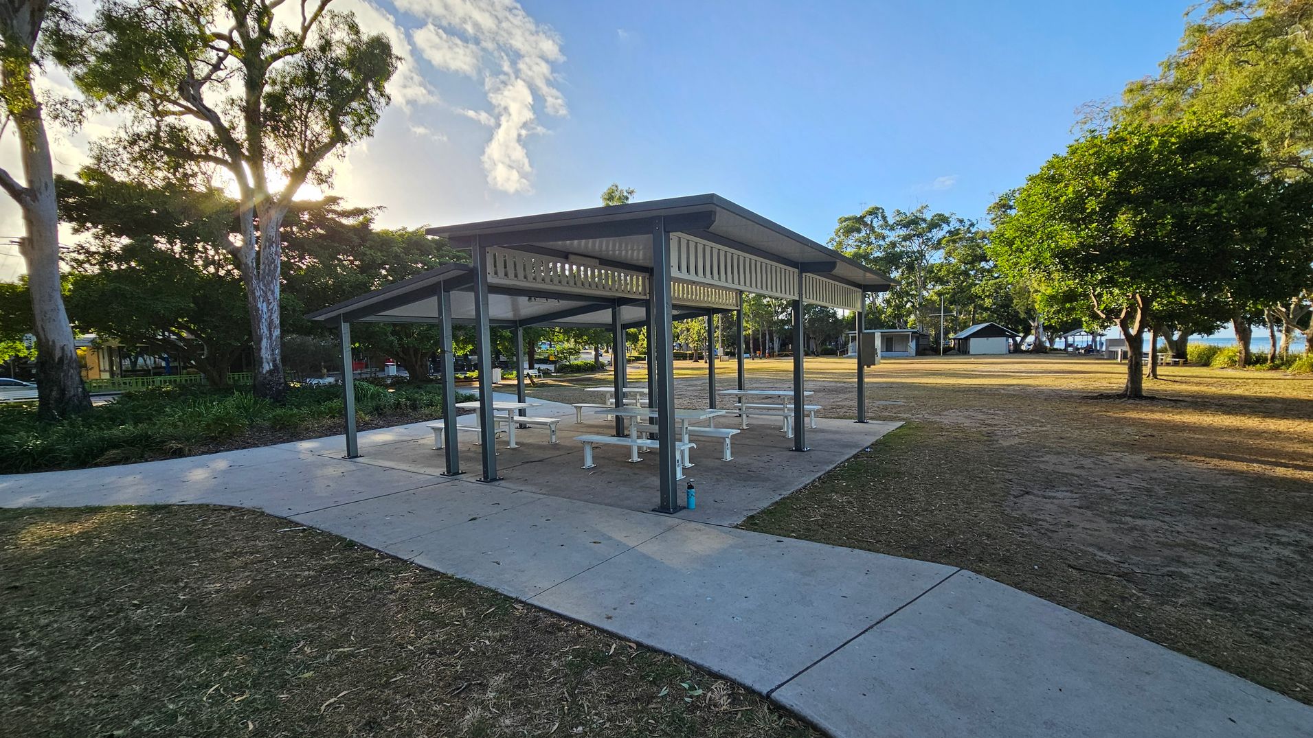 A park setting with a covered picnic area in the centre, featuring several white tables and benches. The structure has a metal roof supported by dark-coloured posts. On the left, tall trees with dense foliage partially block the sun. On the right, more trees and open grassy areas are visible, with a small building in the distance. The ground is a mix of concrete paths and dry grass. A park setting with a covered picnic area in the centre, featuring several white tables and benches. The structure has a metal roof supported by dark-coloured posts. On the left, tall trees with dense foliage partially block the sun. On the right, more trees and open grassy areas are visible, with a small building in the distance. The ground is a mix of concrete paths and dry grass.