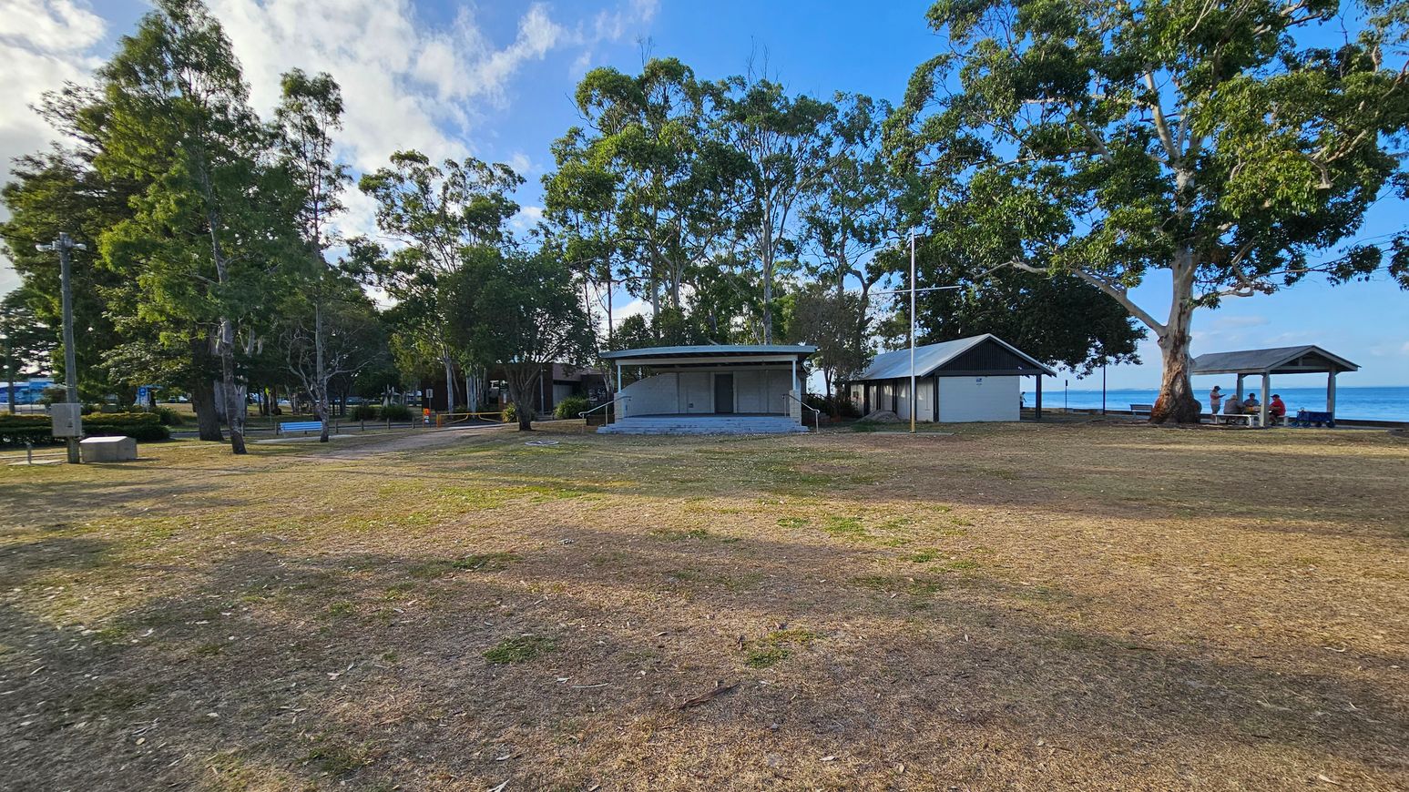 A grassy area with patches of dry soil in the foreground. In the middle, there are two small buildings with metal roofs, surrounded by tall trees. To the right, a group of people sit under a shelter near the water. The background features more trees and a glimpse of the sea. The sky is partly cloudy. A grassy area with patches of dry soil in the foreground. In the middle, there are two small buildings with metal roofs, surrounded by tall trees. To the right, a group of people sit under a shelter near the water. The background features more trees and a glimpse of the sea. The sky is partly cloudy.